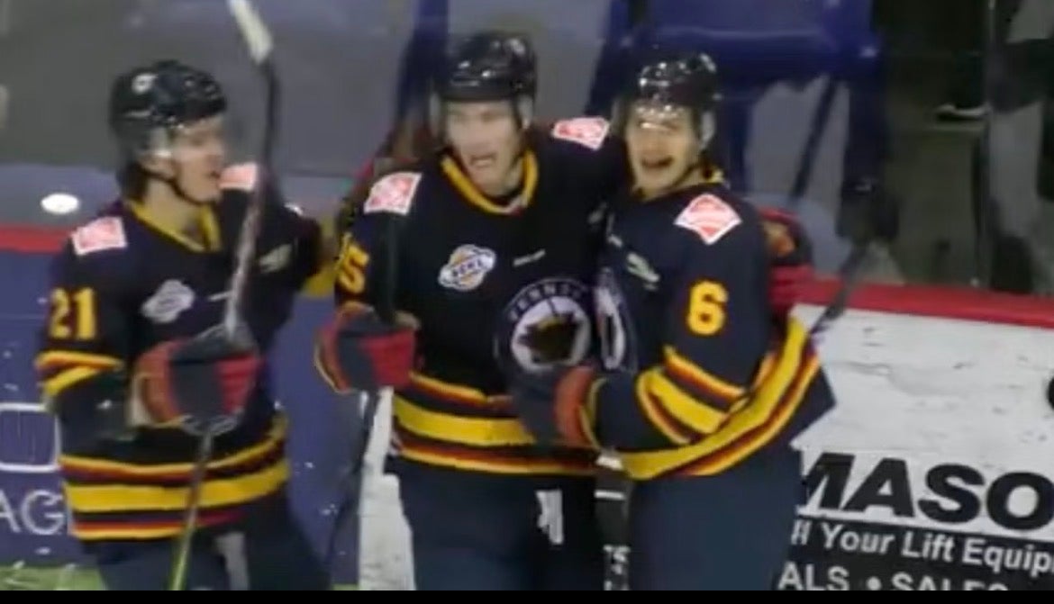Tre ishockeyspelare i svarta tröjor med gula och röda detaljer står tillsammans på isen under en match. De bär hjälmar och håller sina klubbor.