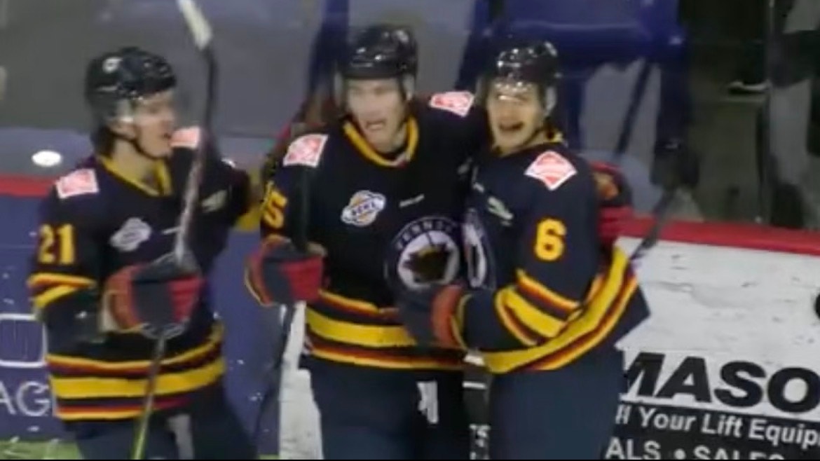Tre ishockeyspelare i svarta tröjor med gula och röda detaljer står tillsammans på isen under en match. De bär hjälmar och håller sina klubbor.