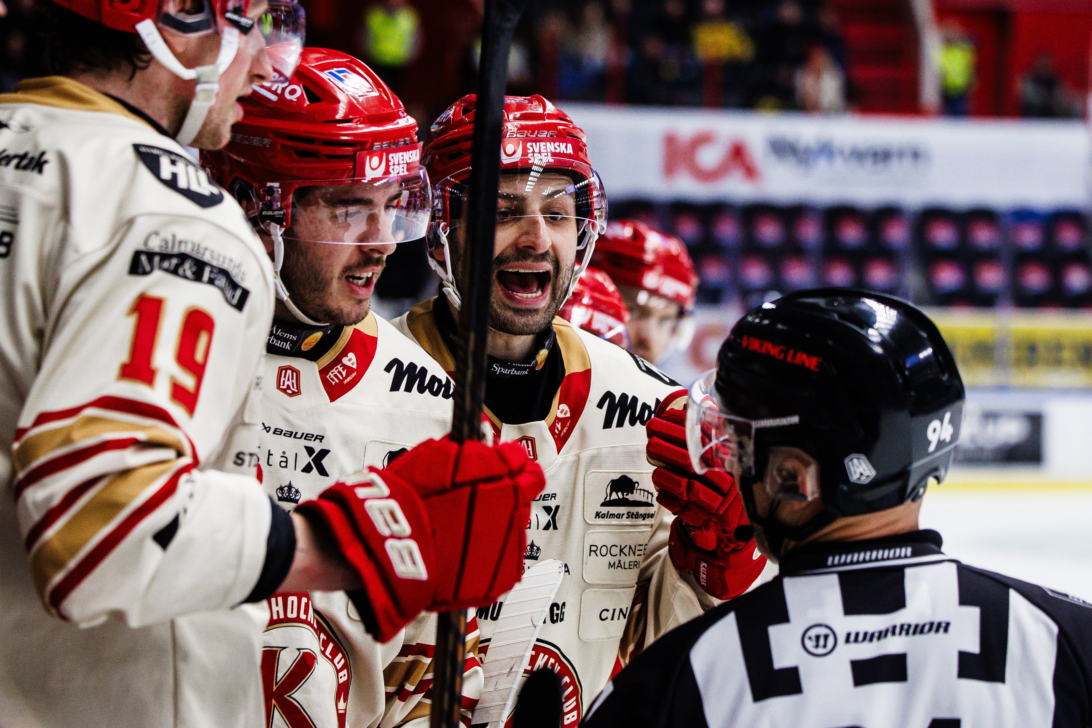 Ishockeymatch där två spelare i röd och vit utrustning diskuterar intensivt med en domare i svart och vit dräkt under en match.
