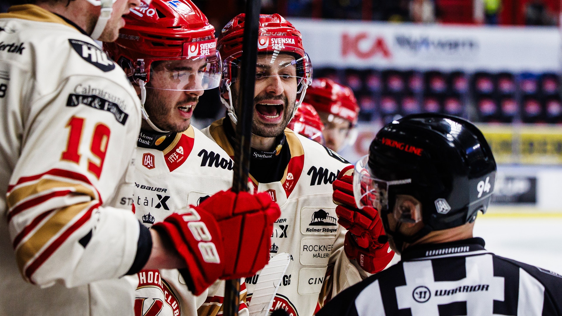 Ishockeymatch där två spelare i röd och vit utrustning diskuterar intensivt med en domare i svart och vit dräkt under en match.