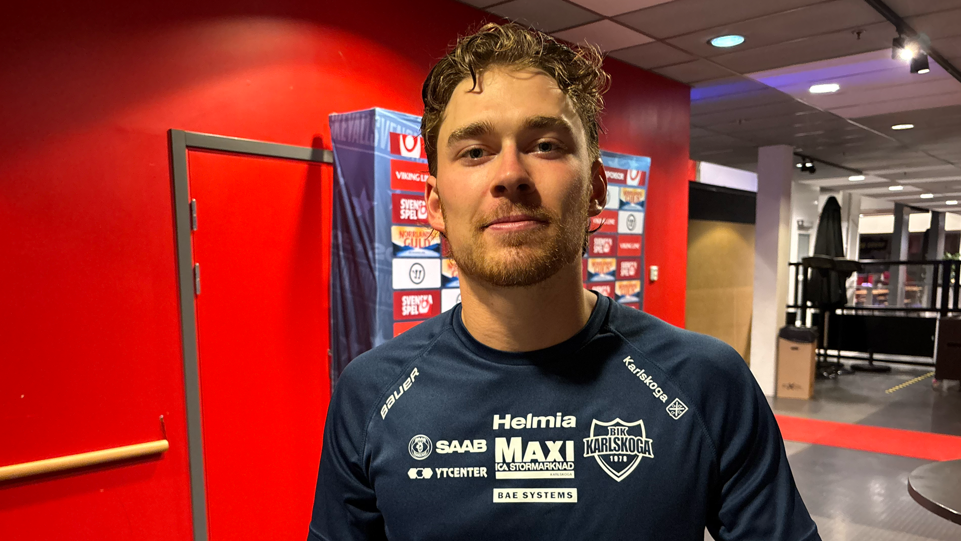 Porträttfoto av en man i en röd sportlokal. Han bär en mörkblå tröja med sponsorlogotyper och står framför en röd vägg med olika emblemer.