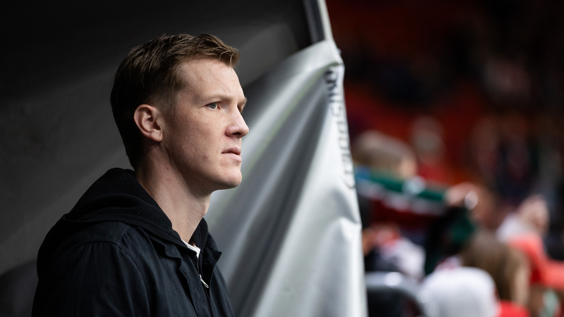 Man i svart jacka vid ishockeybåset tittar ut över isen under en match.