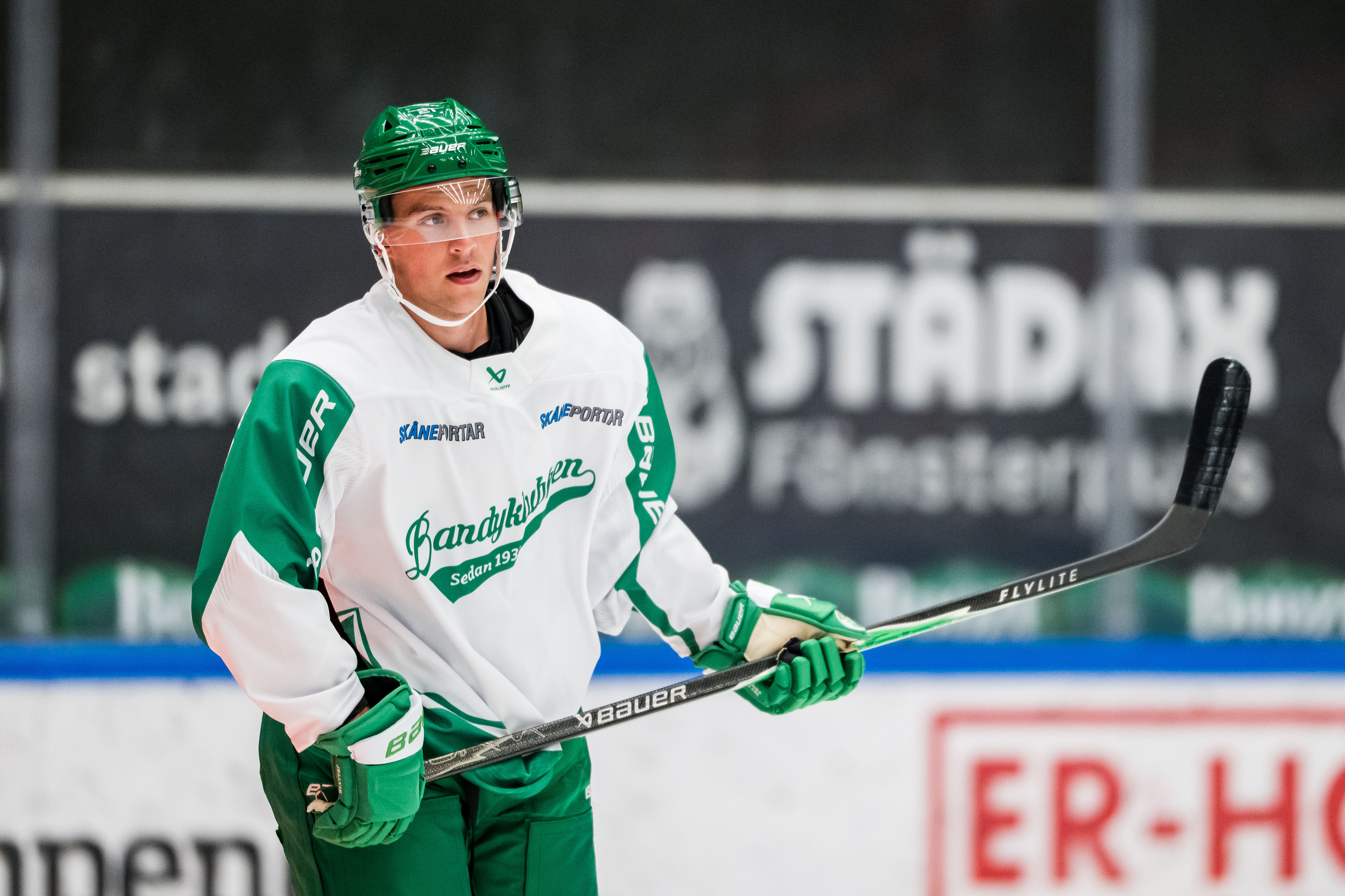 Ishockeyspelare i grön och vit matchdräkt som står på isen med hockeyklubbe i händerna.