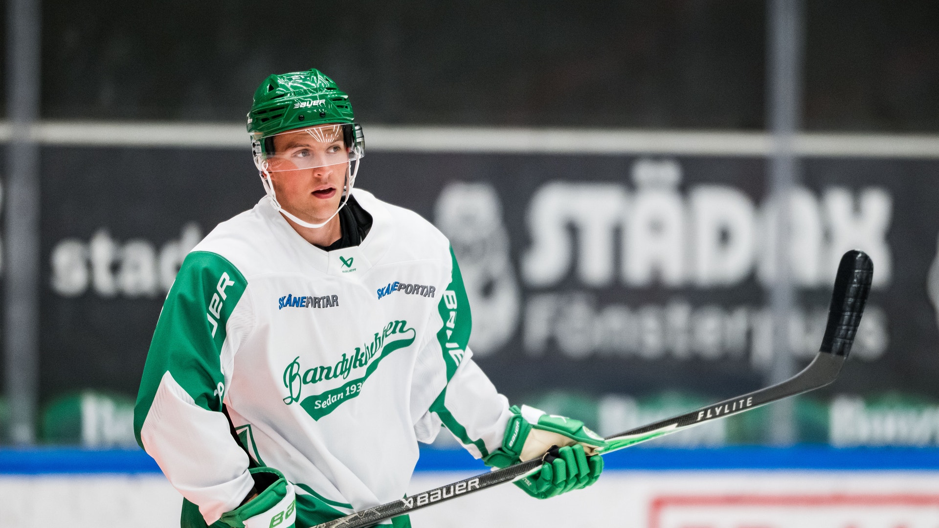 Ishockeyspelare i grön och vit matchdräkt som står på isen med hockeyklubbe i händerna.