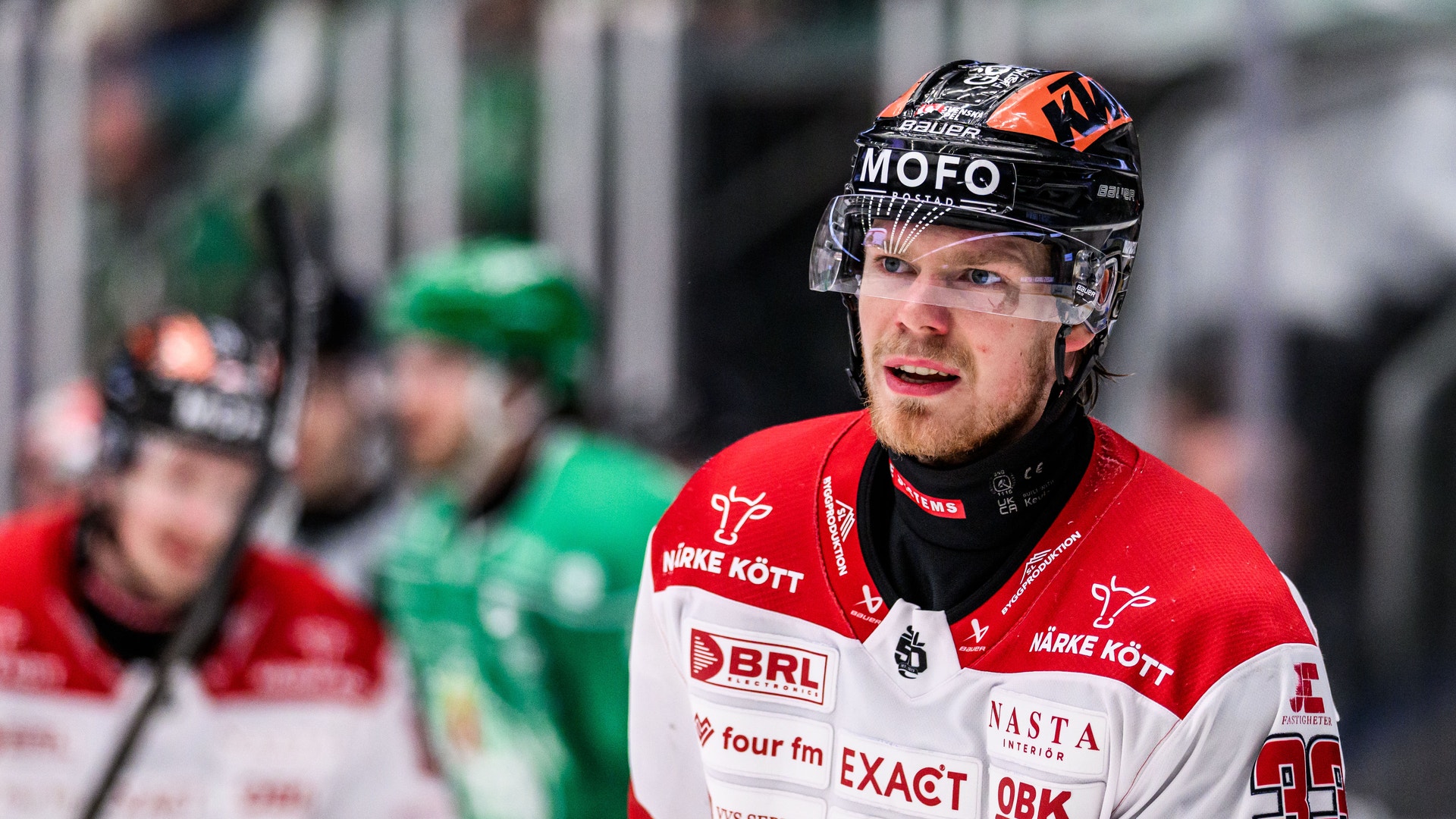 Ishockeyspelare i röd och vit matchdräkt under en match. Spelaren bär hjälm med visir och är omgiven av andra spelare på isen.