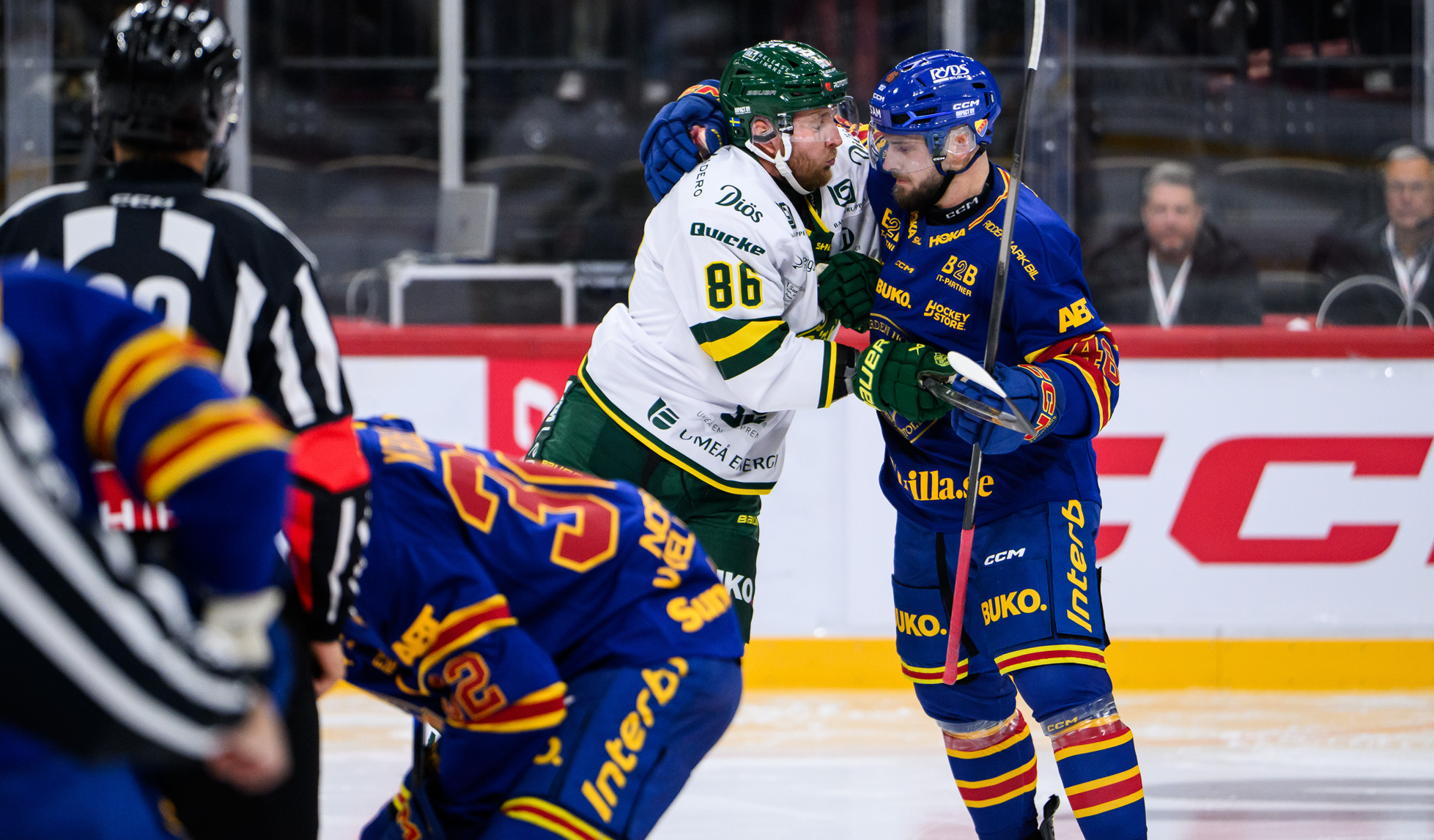 Två ishockeyspelare från olika lag knuffas och tjafsar med varandra på isen, medan en domare observerar.