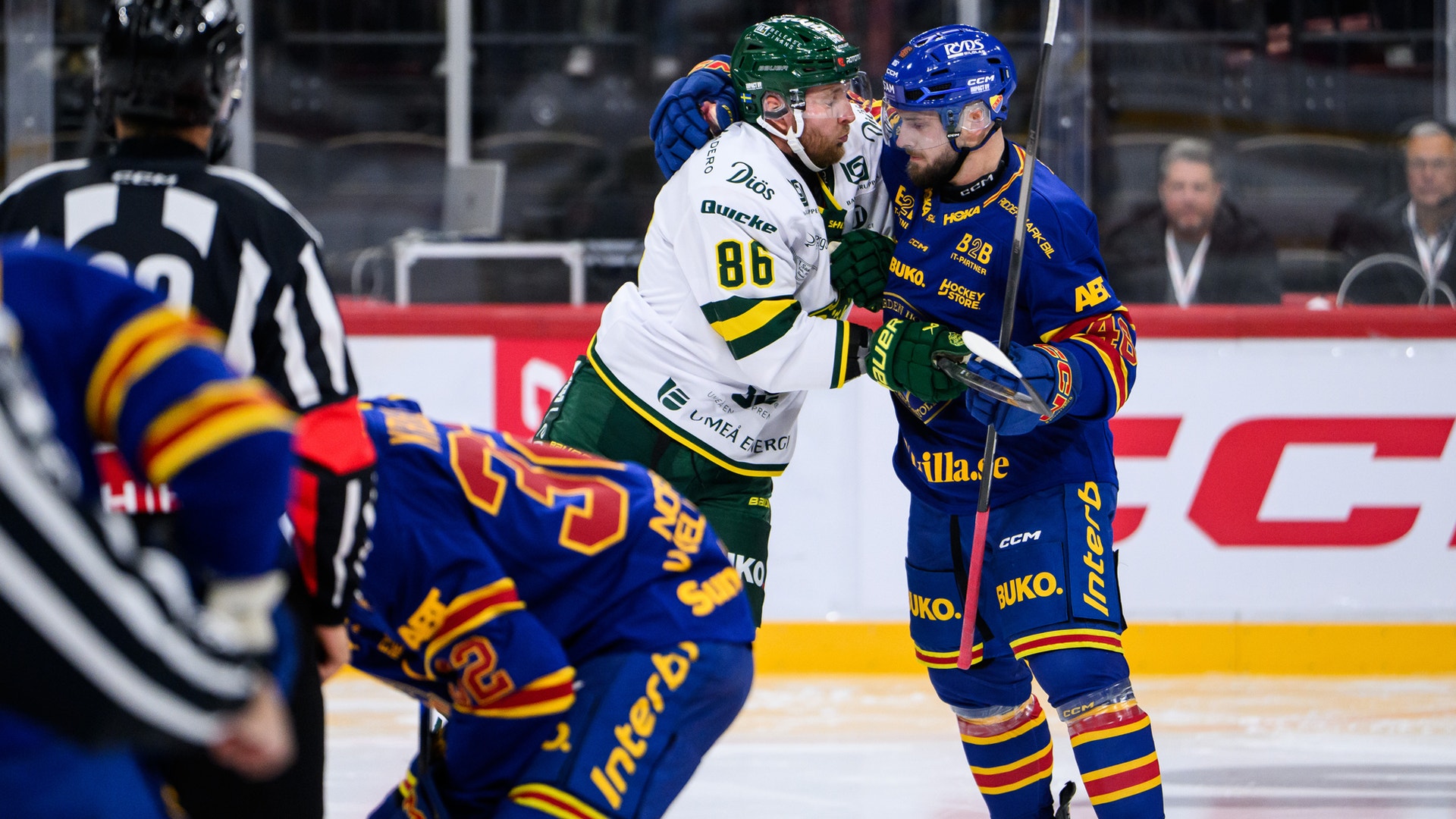 Två ishockeyspelare från olika lag knuffas och tjafsar med varandra på isen, medan en domare observerar.
