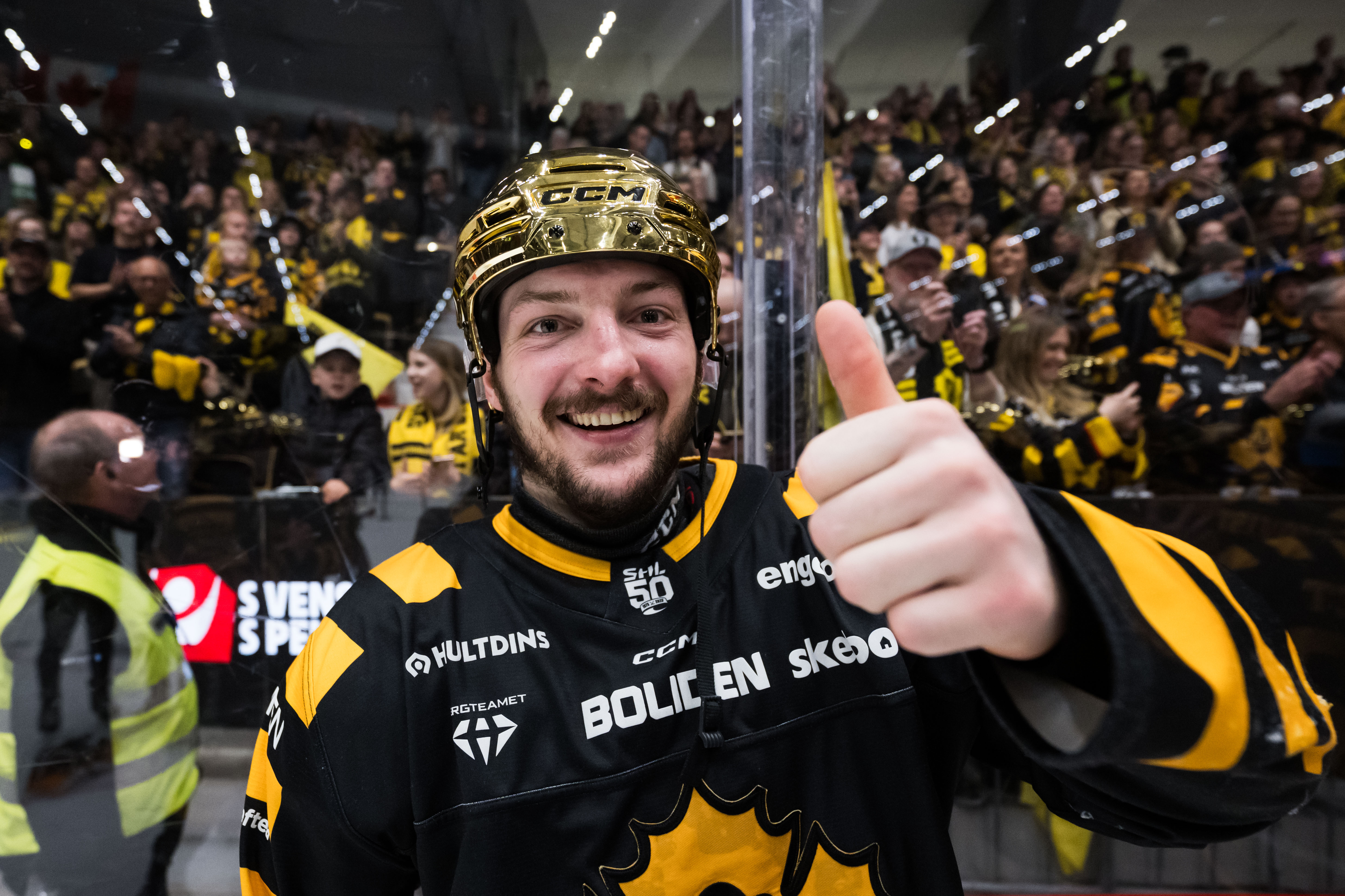 Ishockeyspelare i svart och gul uniform ger tummen upp framför en fullsatt arena med supportrar.
