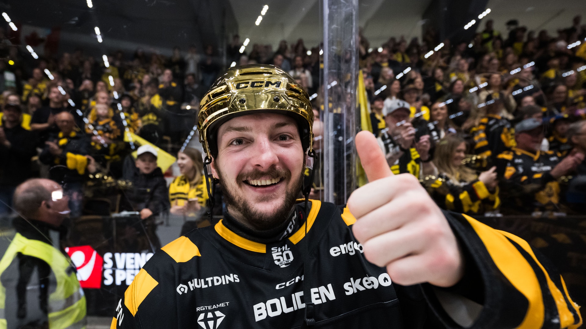 Ishockeyspelare i svart och gul uniform ger tummen upp framför en fullsatt arena med supportrar.