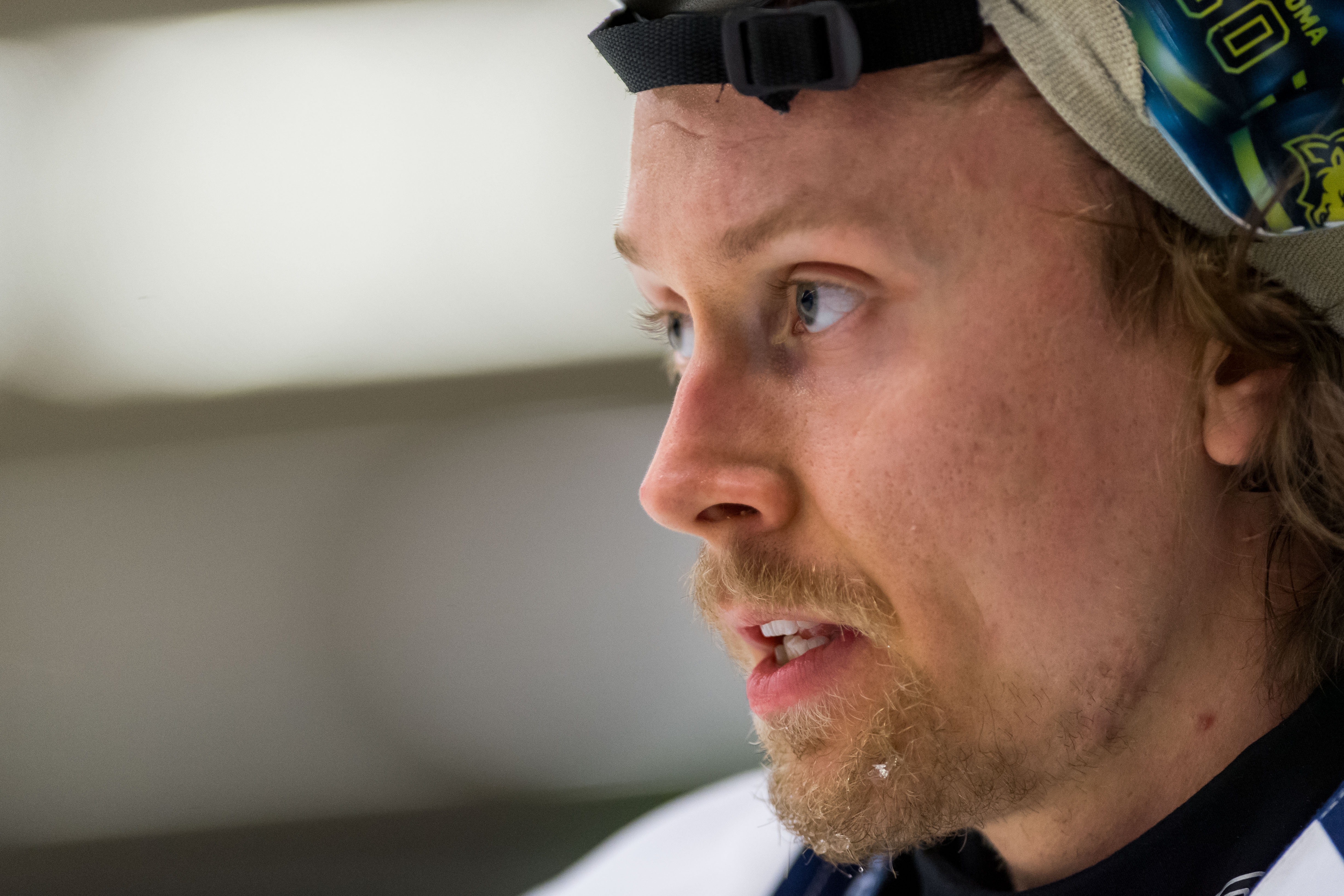 Man i profil med mörkblå keps och sportkläder. Han har rödbrunt hår och skägg. Porträtt av en man i svart kavaj och mörkblå tröja, fotograferad inomhus. I bakgrunden syns en gul och blå banderoll och andra personer.