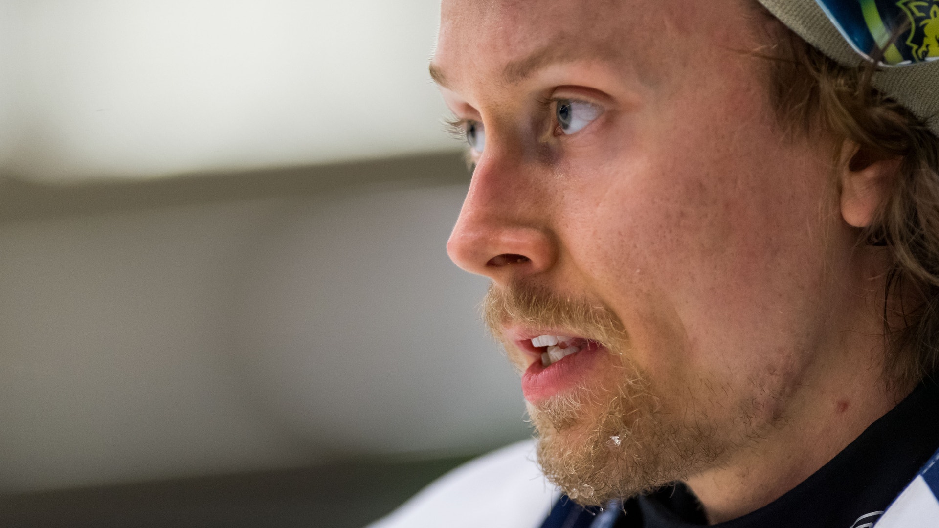 Man i profil med mörkblå keps och sportkläder. Han har rödbrunt hår och skägg. Porträtt av en man i svart kavaj och mörkblå tröja, fotograferad inomhus. I bakgrunden syns en gul och blå banderoll och andra personer.