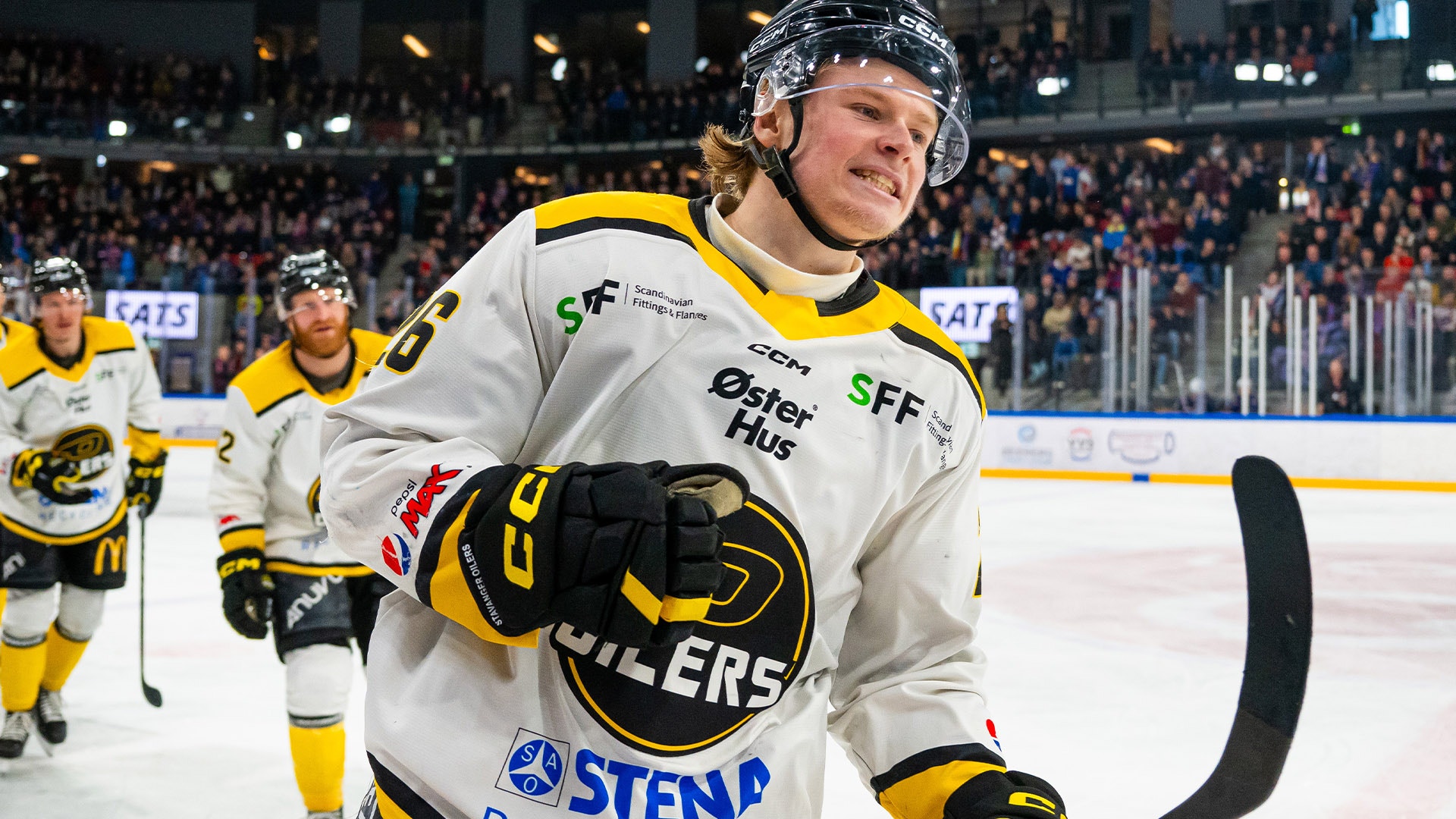Ishockeyspelare i gul och vit uniform poserar på isen under en match med fullsatt läktare i bakgrunden.