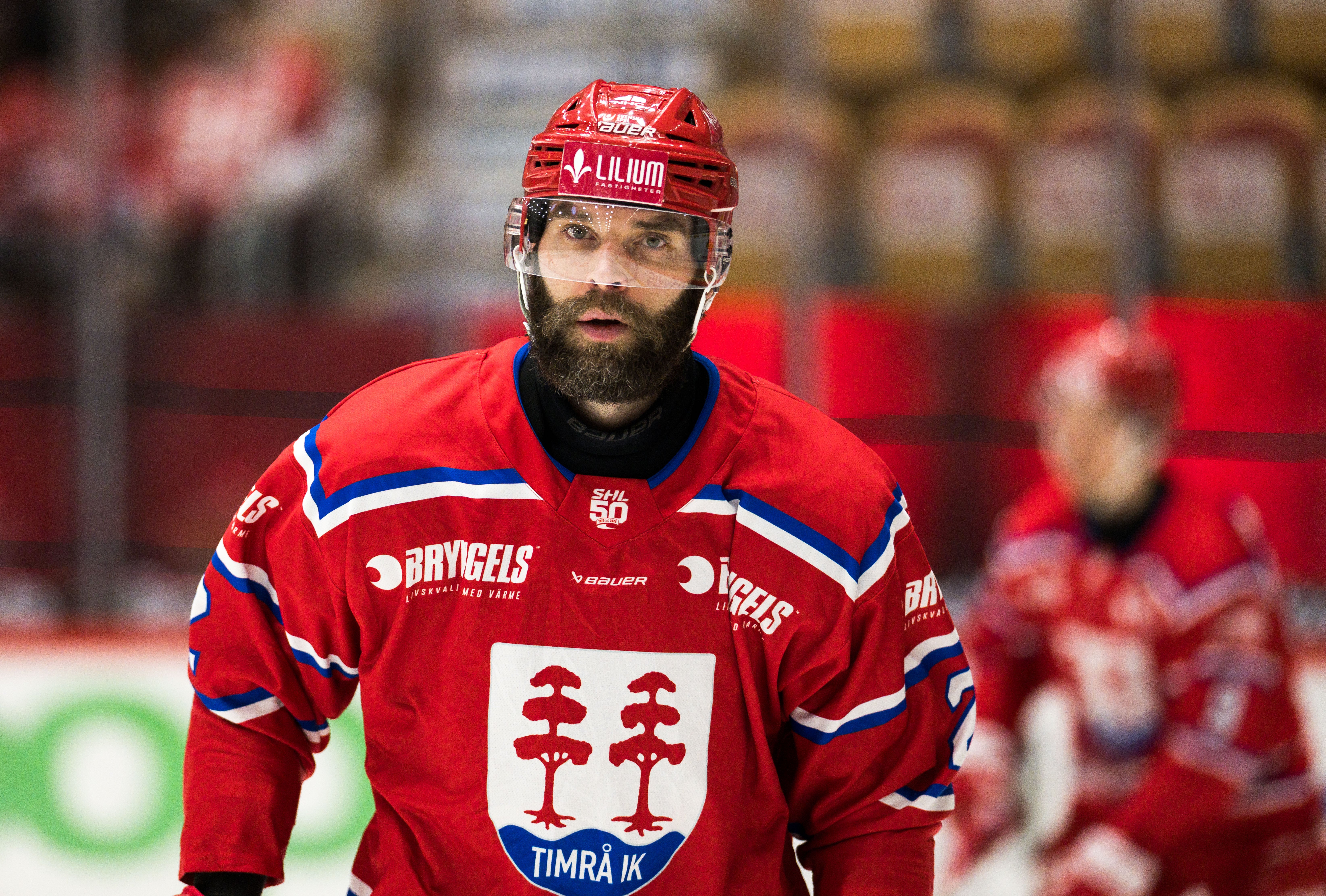 Ishockeyspelare i röd landstruppa med hjälm och glasögon under en ishockeymatch. Porträtt av en man med glasögon och skägg, iklädd en mörkblå jacka mot en suddig röd bakgrund.