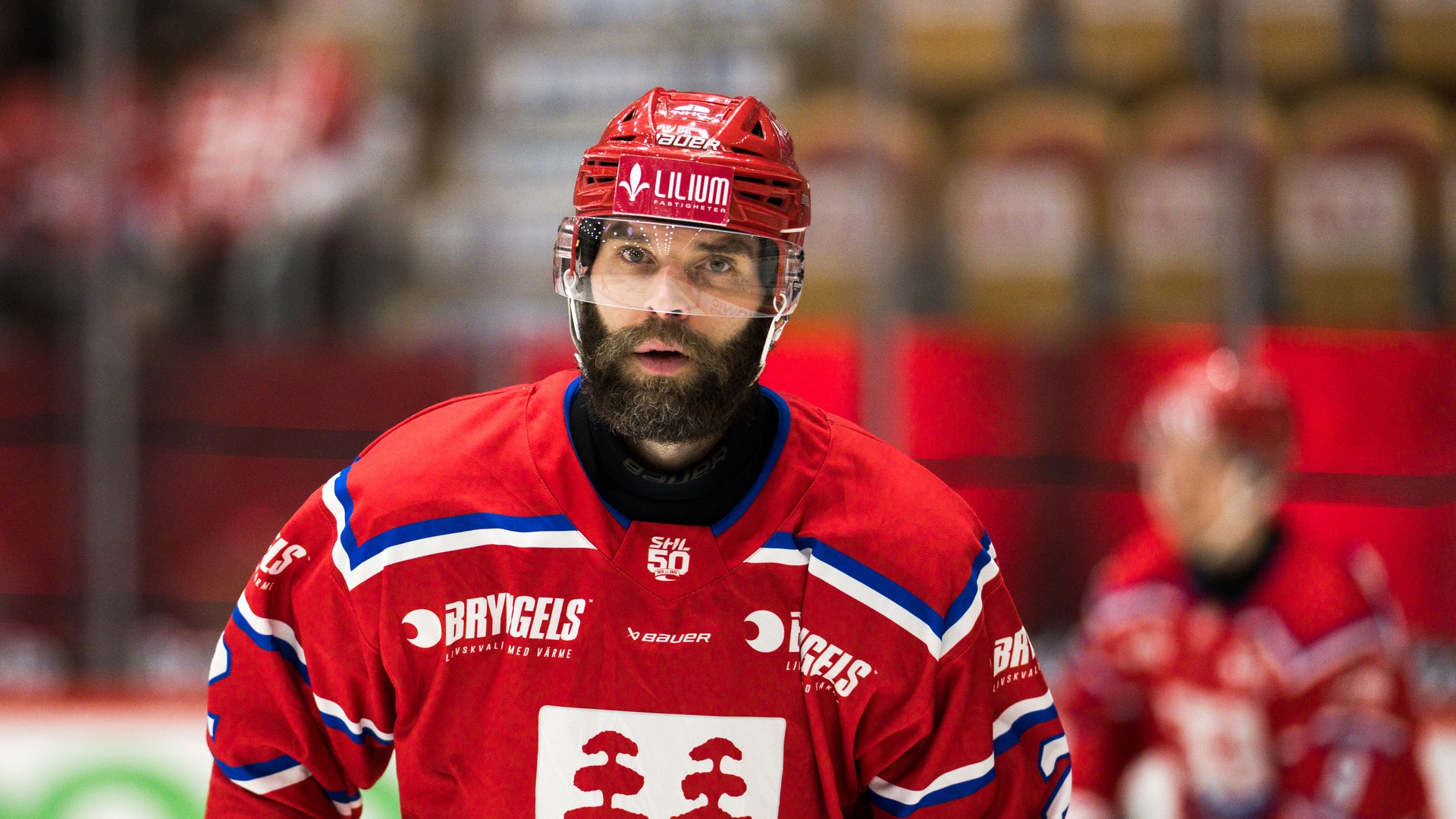 Ishockeyspelare i röd landstruppa med hjälm och glasögon under en ishockeymatch. Porträtt av en man med glasögon och skägg, iklädd en mörkblå jacka mot en suddig röd bakgrund.