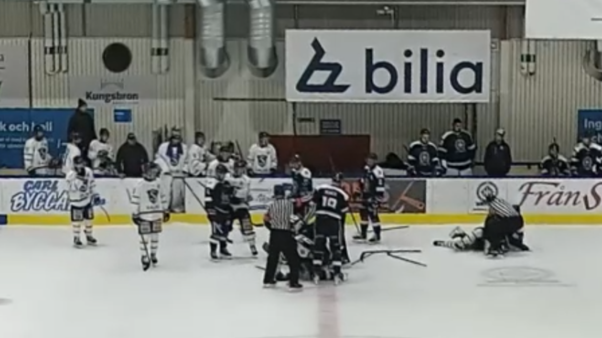Ishockeyspelare från två lag samlade på isen i en hall, där en domare står mitt i gruppen och flera spelare ligger på isen. Spelare och ledare tittar på från sidlinjen.