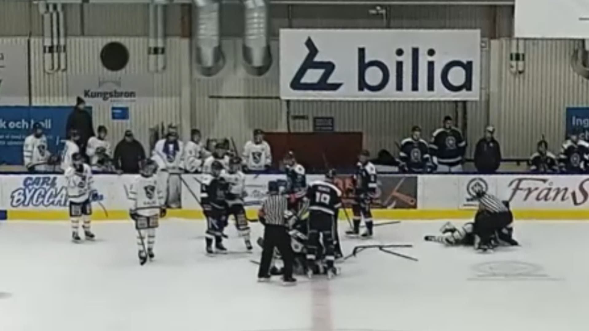 Ishockeyspelare från två lag samlade på isen i en hall, där en domare står mitt i gruppen och flera spelare ligger på isen. Spelare och ledare tittar på från sidlinjen.