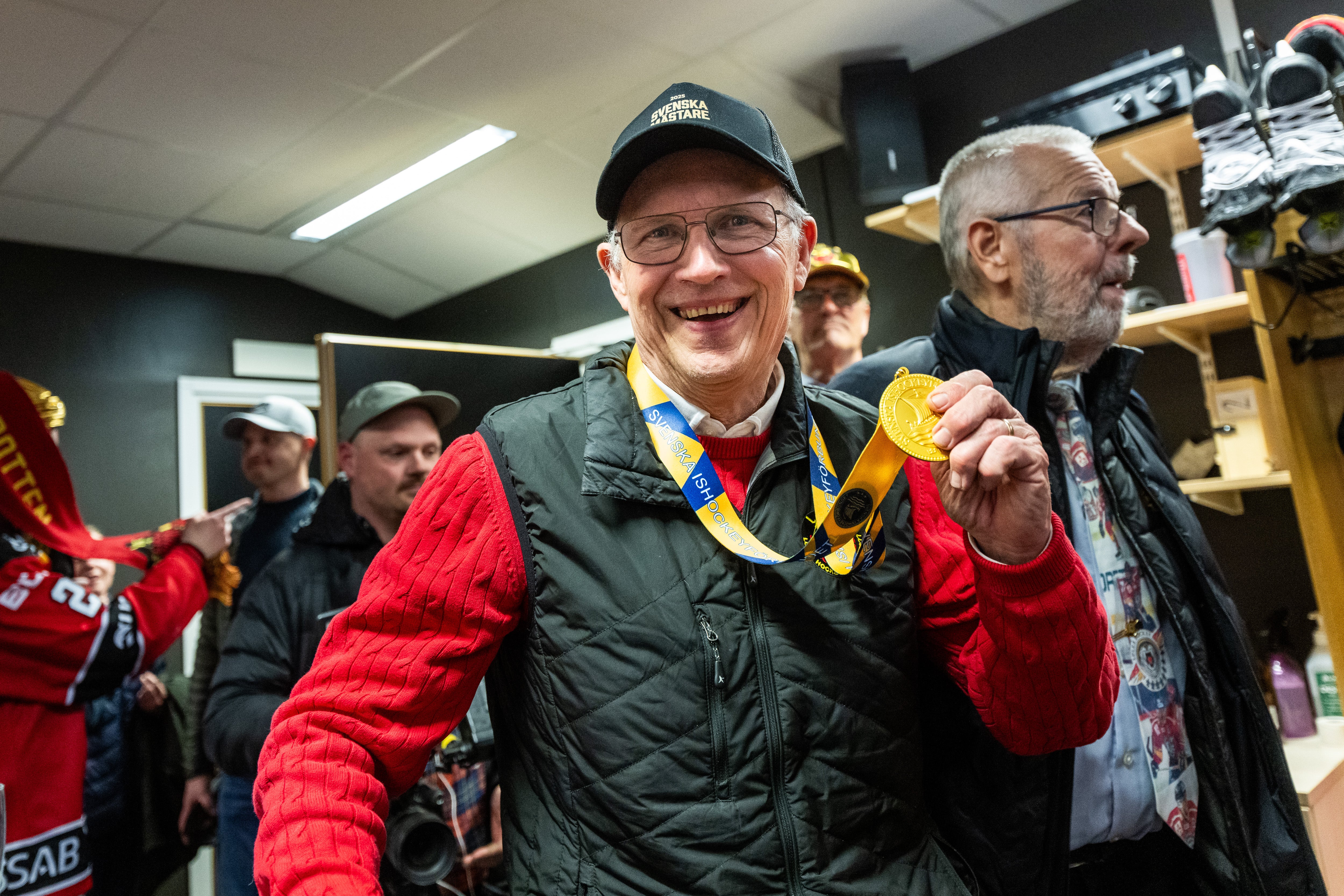 En man i röd och svart jacka med glasögon och keps visar stolt upp en guldmedalj runt halsen. Han står tillsammans med andra personer inomhus och ler mot kameran.