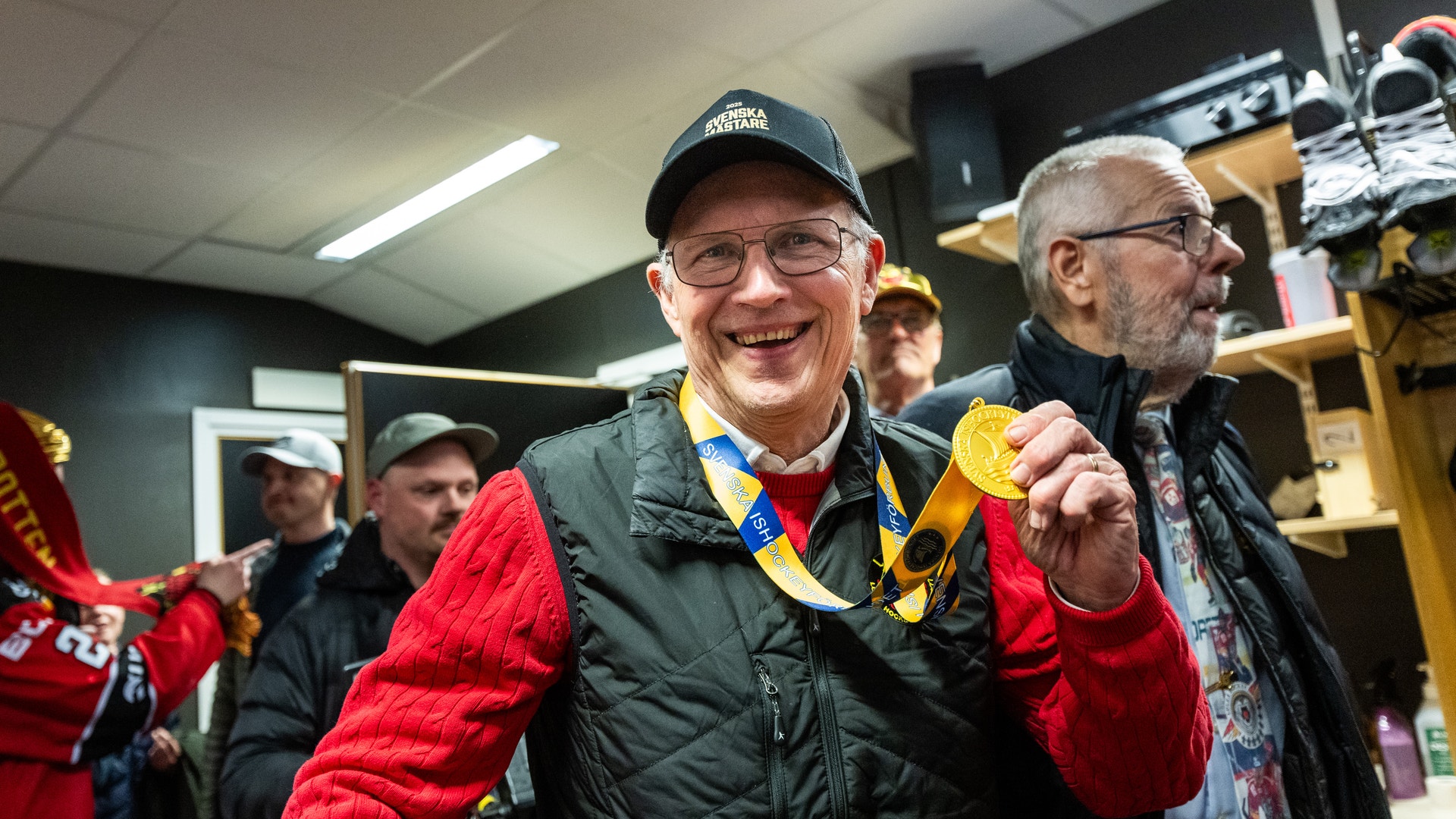 En man i röd och svart jacka med glasögon och keps visar stolt upp en guldmedalj runt halsen. Han står tillsammans med andra personer inomhus och ler mot kameran.