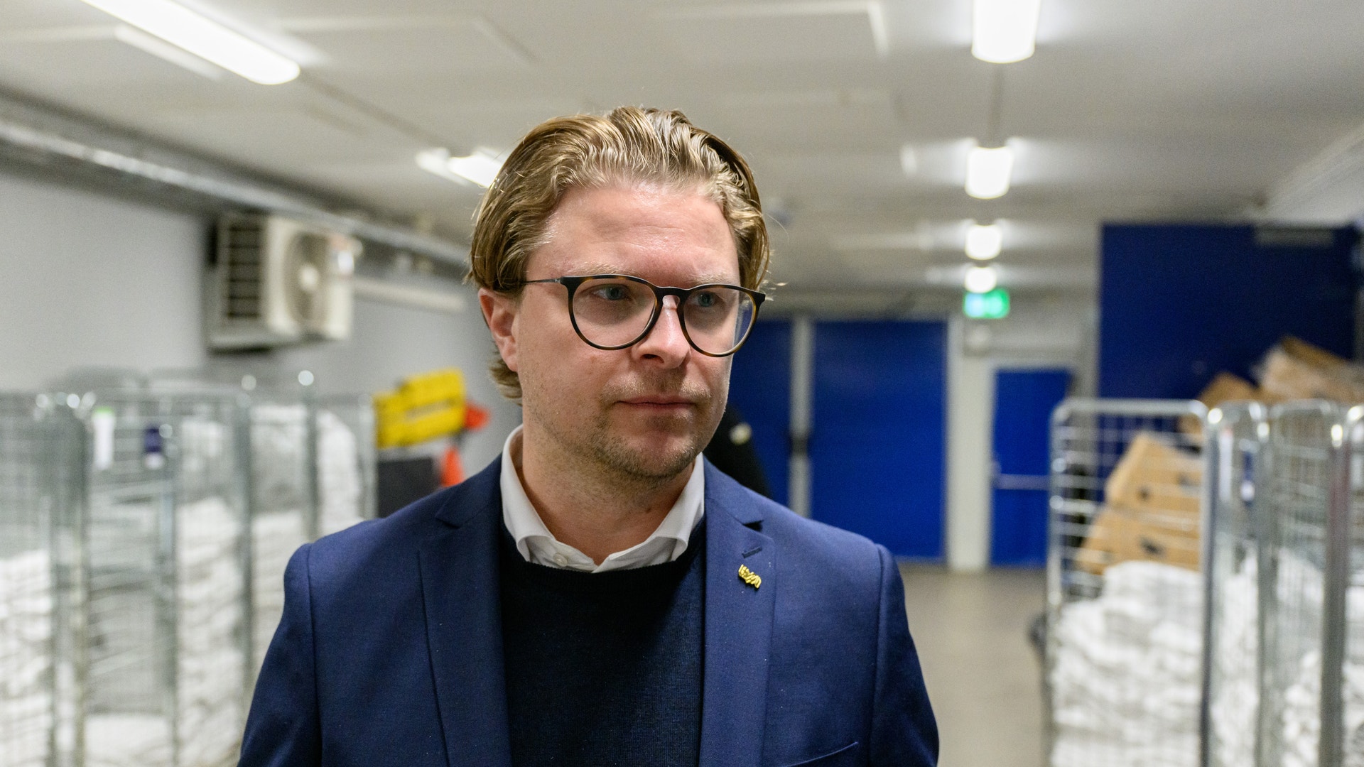 En man med glasögon och blå blazer står i ett lager eller industrianläggning med blå dörrar och lagringsutrustning i bakgrunden.