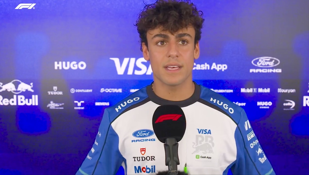 En ung F1-förare i racingoverall intervjuas framför en mikrofon med sponsorlogotyper i bakgrunden.
