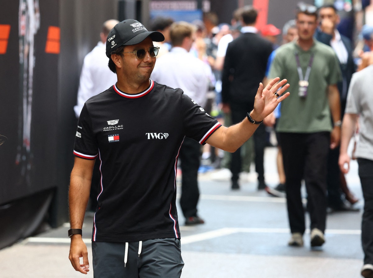 Formel 1-förare i svart teamkläder går genom paddocken på en racingklubb. Han bär solglasögon och svart keps och gestikulerar medan han går förbi andra människor.