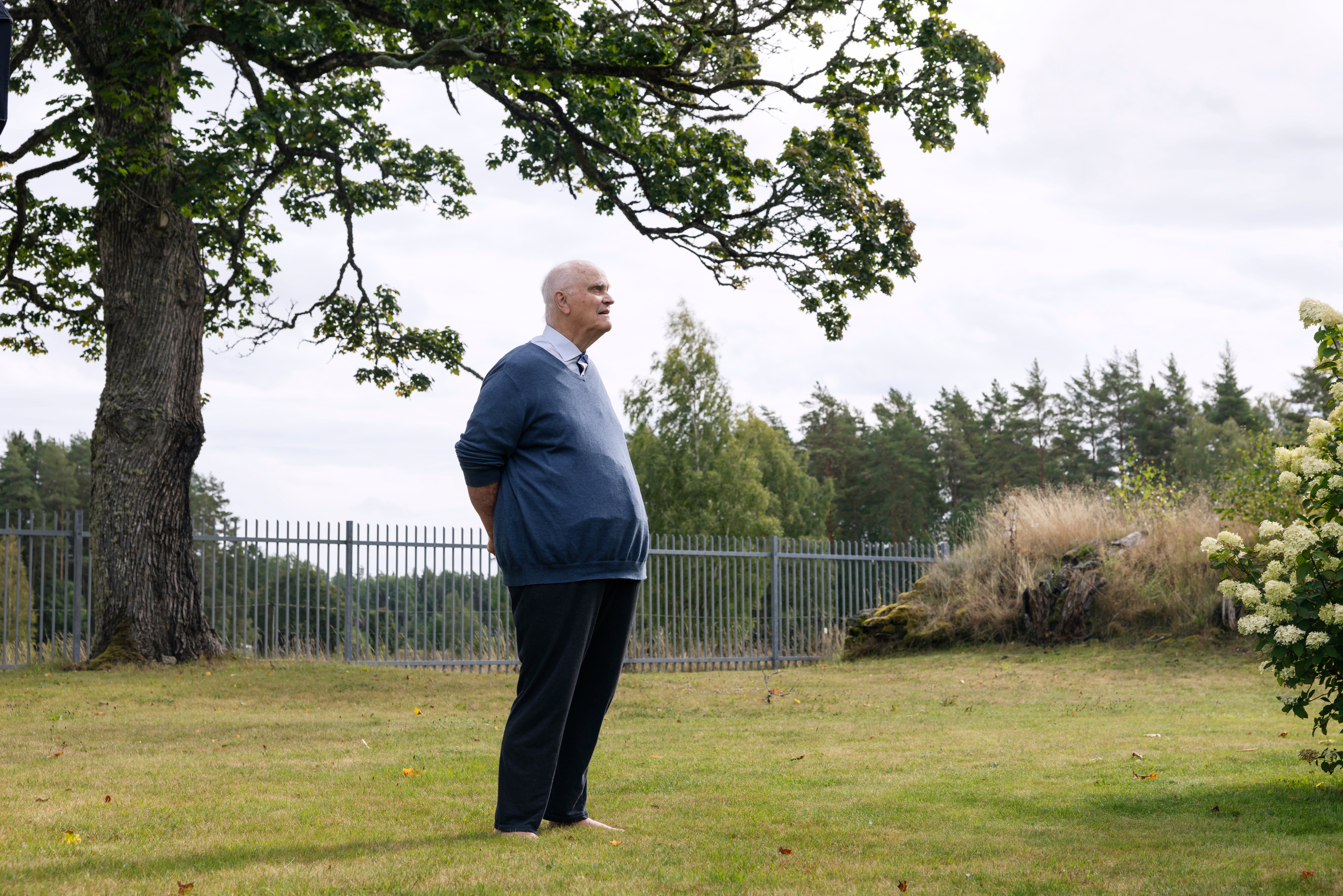 En man i blå jacka står på en grässmatta vid ett stort träd. I bakgrunden syns ett vitt staket och skog.