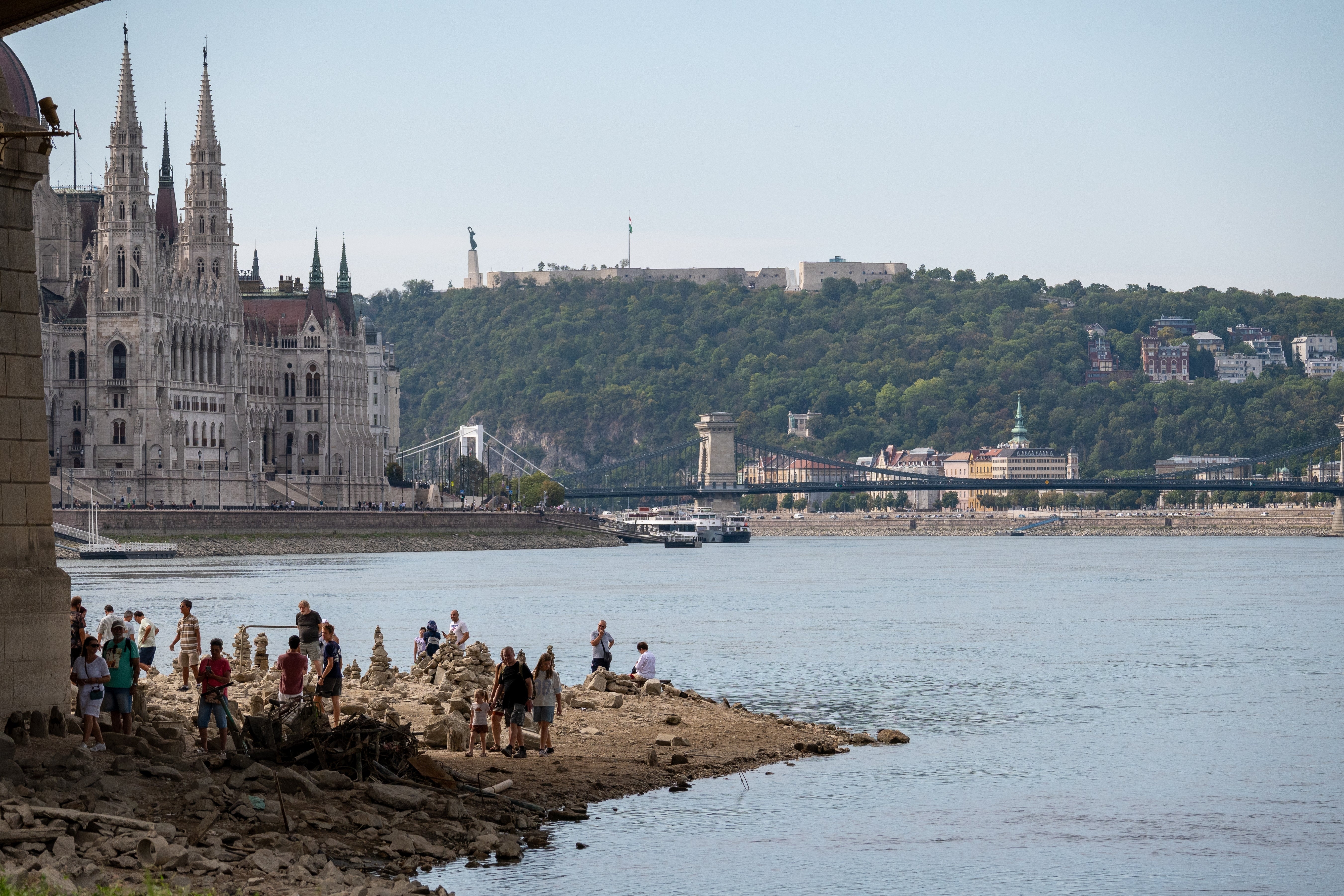 Utsikt över Donau i Budapest med parlamentsbyggnaden och en bro i bakgrunden, och människor som vistas vid flodstranden.
