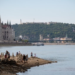 Utsikt över Donau i Budapest med parlamentsbyggnaden och en bro i bakgrunden, och människor som vistas vid flodstranden.
