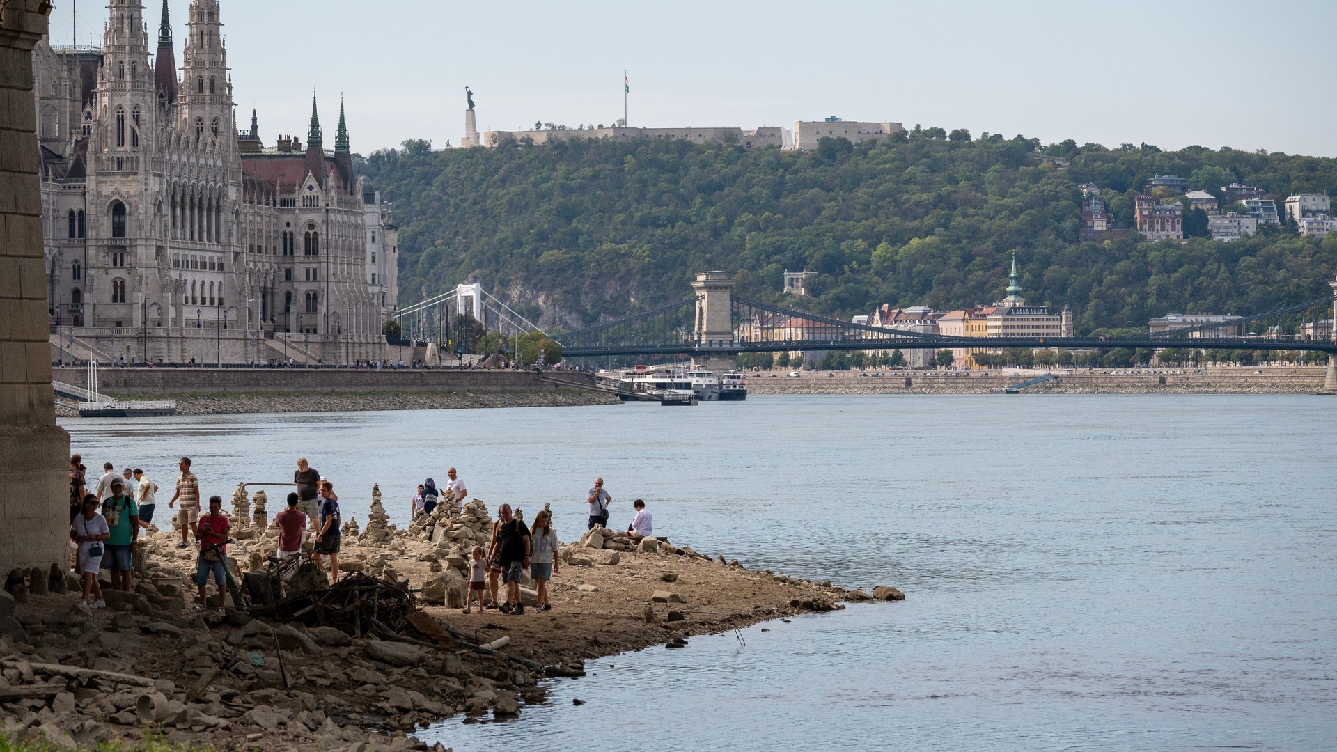 Utsikt över Donau i Budapest med parlamentsbyggnaden och en bro i bakgrunden, och människor som vistas vid flodstranden.