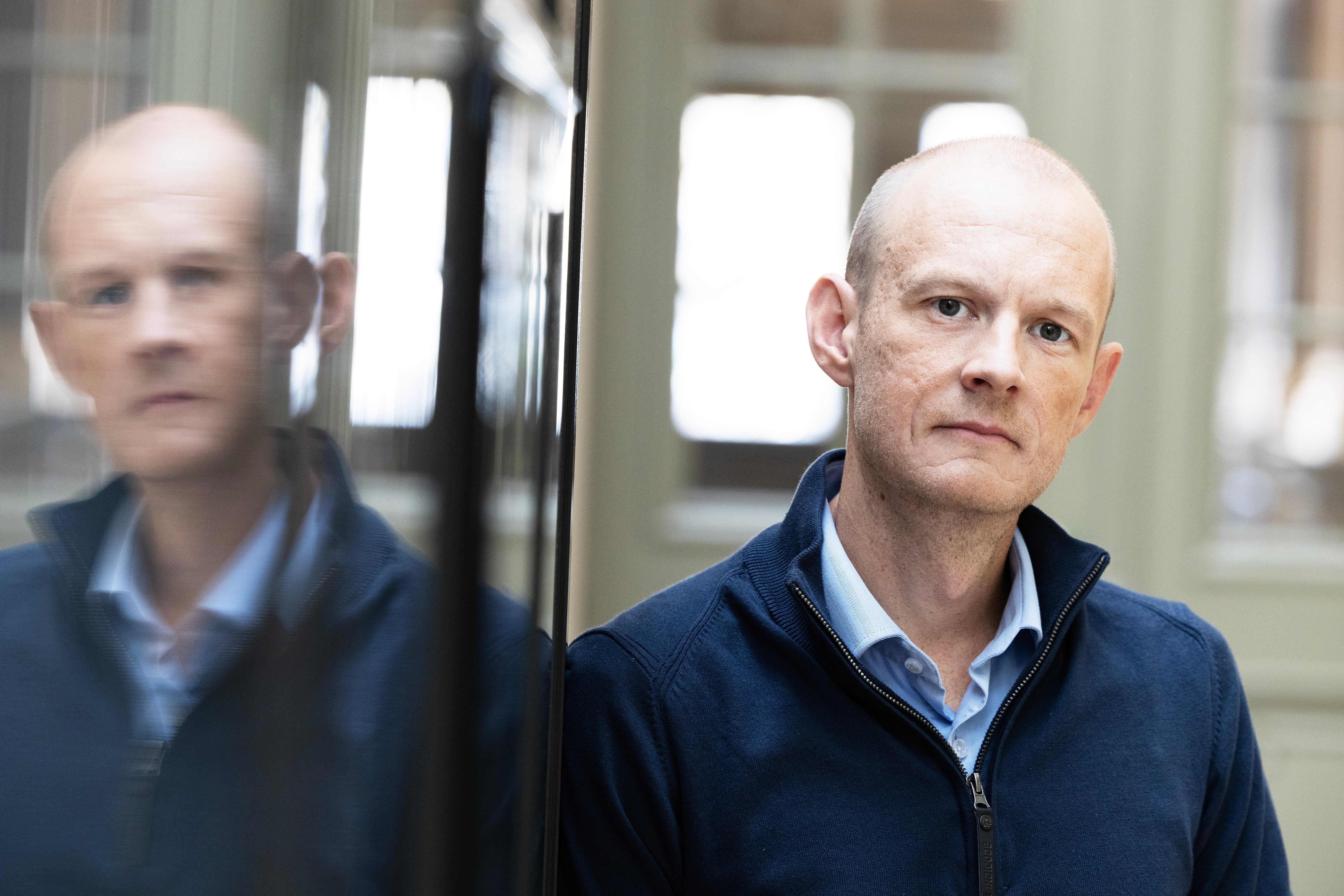 Porträtt av en man i mörkblå jacka framför en glasvägg som reflekterar hans bild.