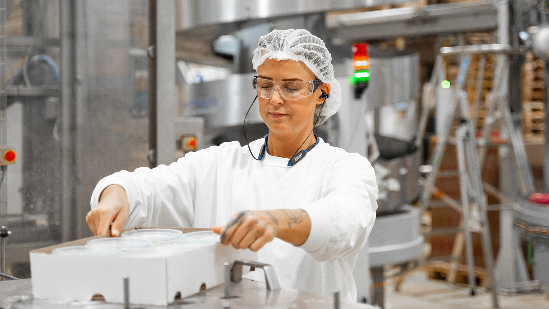 Arbetare i vita skyddskläder hanterar en behållare i en industriell produktionsmiljö.