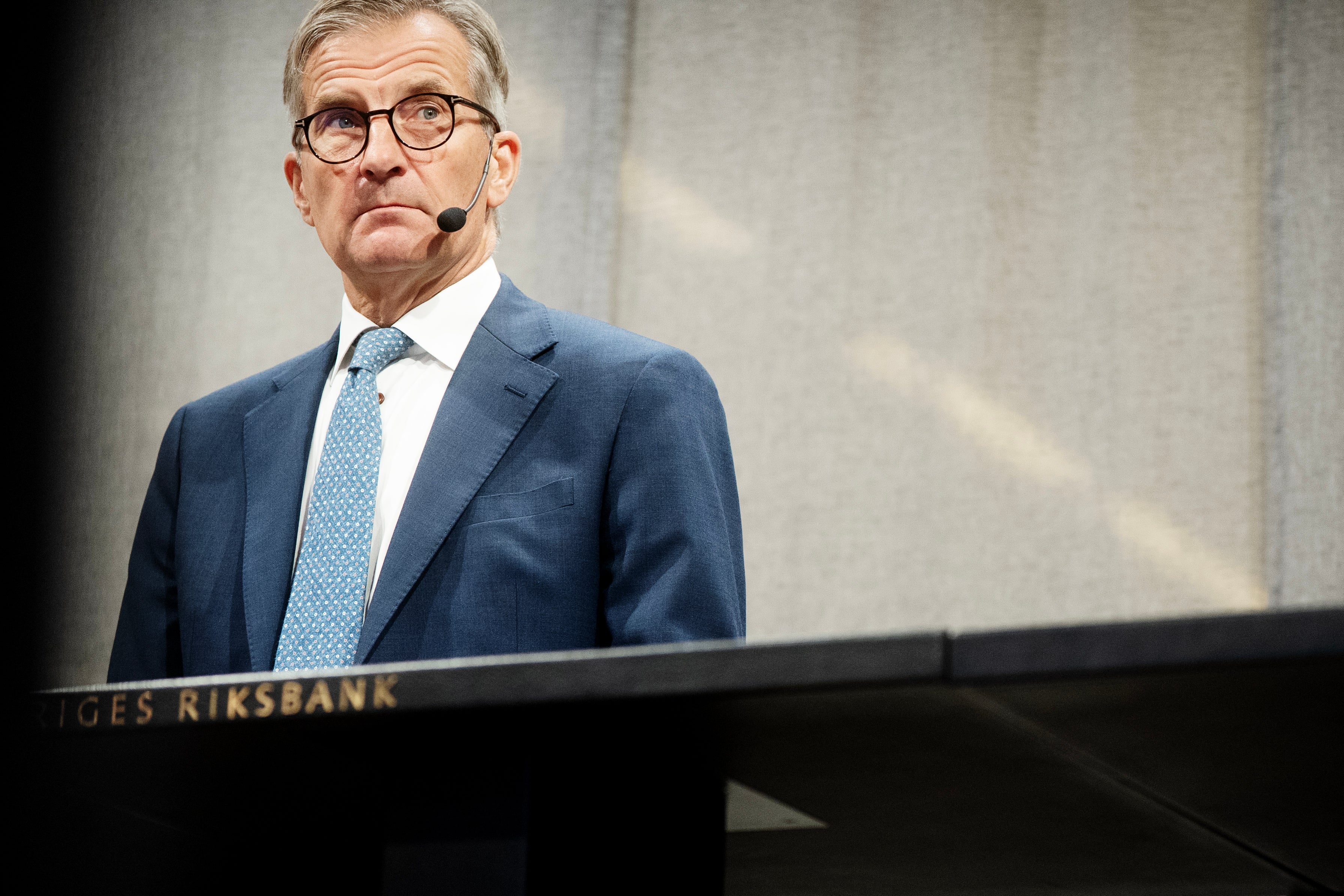 En man i blå kostym och glasögon står vid ett podium märkt Sveriges Riksbank och håller ett tal med headset-mikrofon.