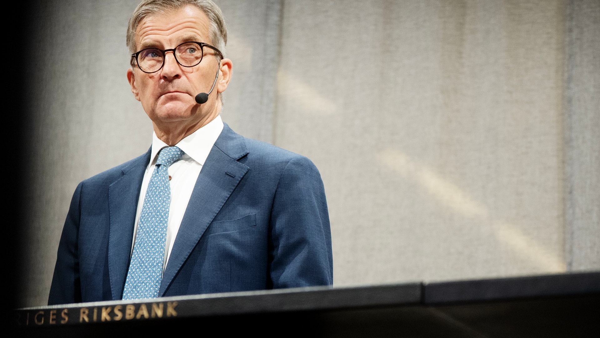 En man i blå kostym och glasögon står vid ett podium märkt Sveriges Riksbank och håller ett tal med headset-mikrofon.