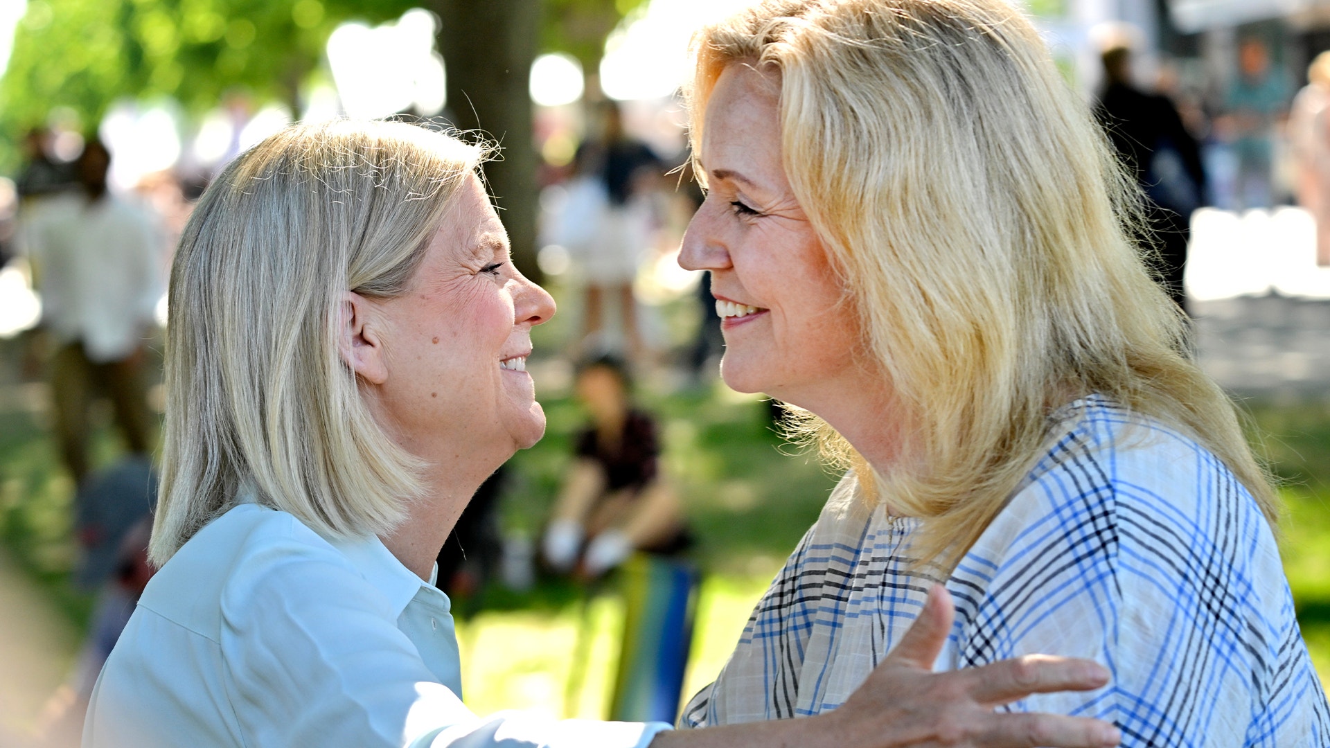 Två kvinnor ler och kramar varandra utomhus i en park.