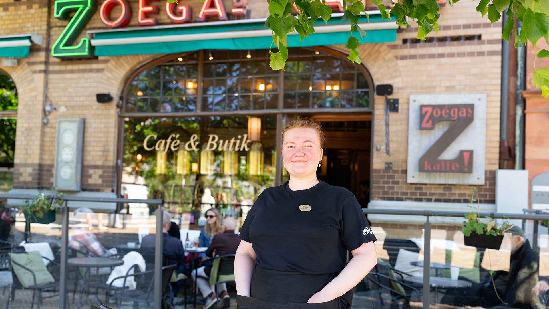 Emelie Gestrén, platschef på Café 1886 Helsingborg, står framför caféets entré och ler mot kameran. Hon är klädd i svart och caféets fasad med skyltfönster syns i bakgrunden.