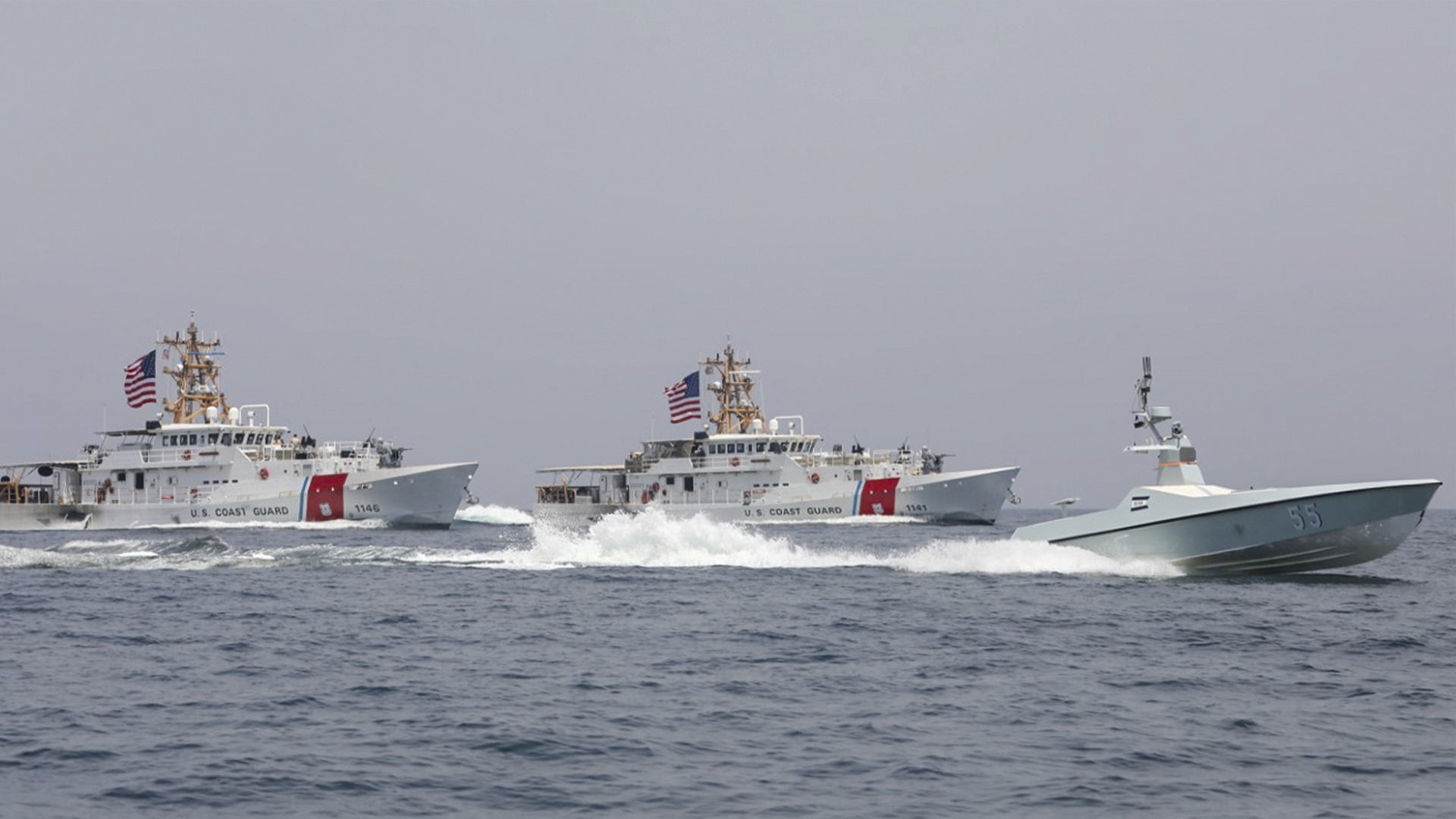 Tre militära fartyg på havet, två större grå fartyg med amerikanska flaggor och ett mindre vitt fartyg.