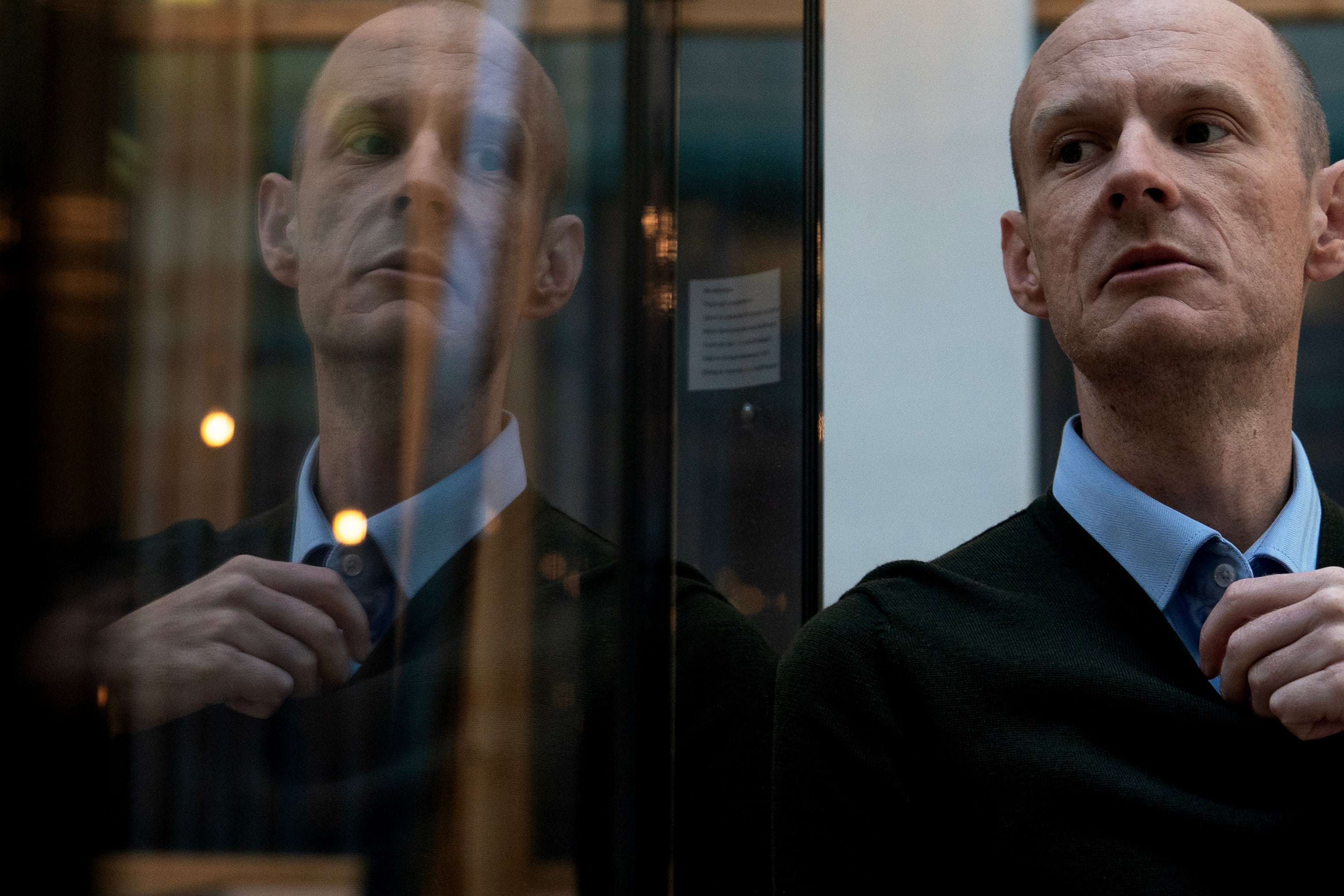 En flintskallig man i blå skjorta och mörk tröja blickar åt sidan. Hans spegelbild syns i en glasyta bredvid honom.