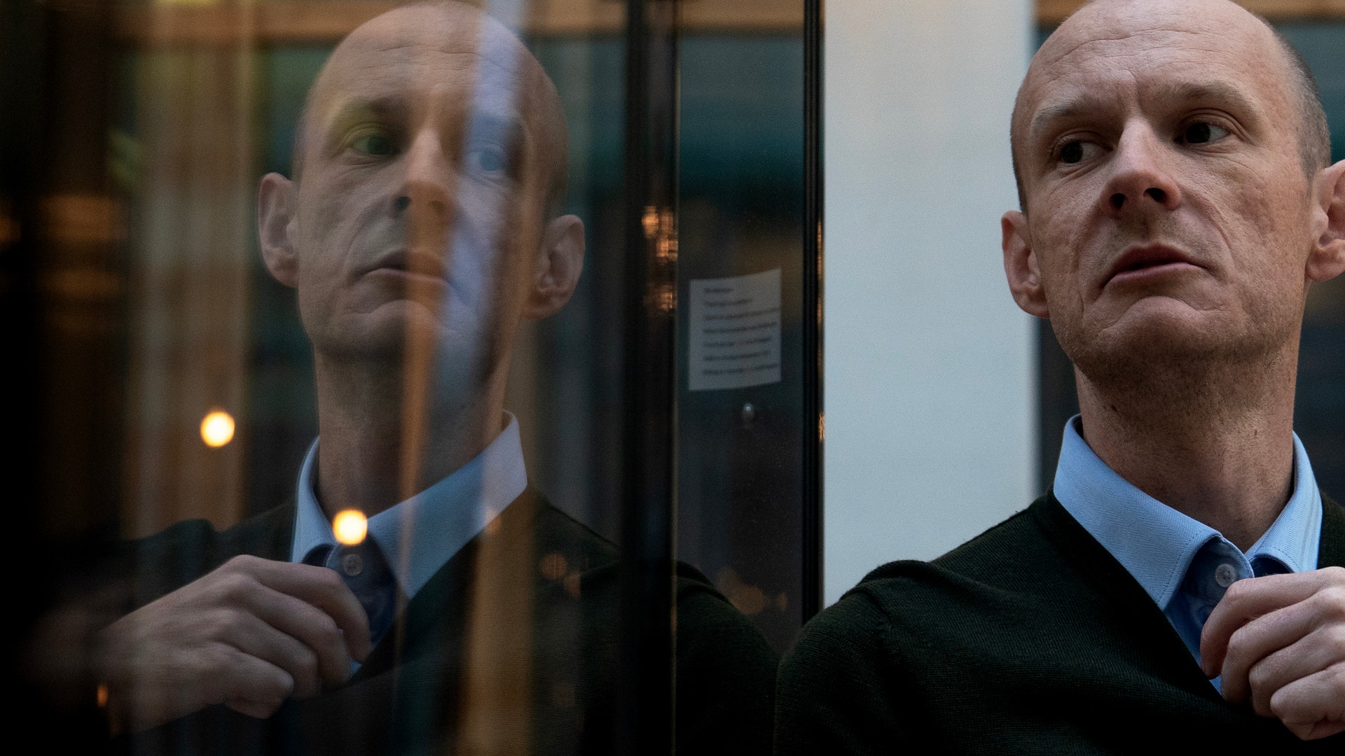 En flintskallig man i blå skjorta och mörk tröja blickar åt sidan. Hans spegelbild syns i en glasyta bredvid honom.