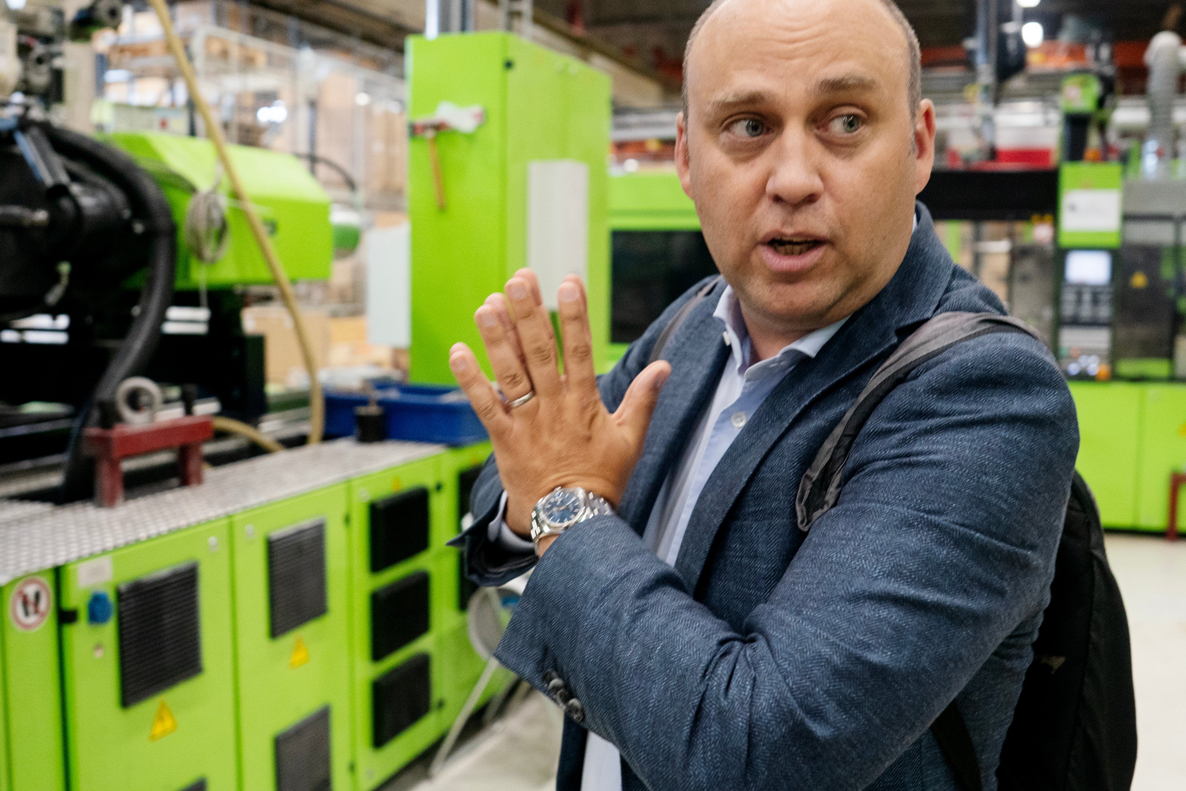 En man i mörkblå jacka gestikulerar och talar i en industriell miljö omgiven av grön maskinutrustning.