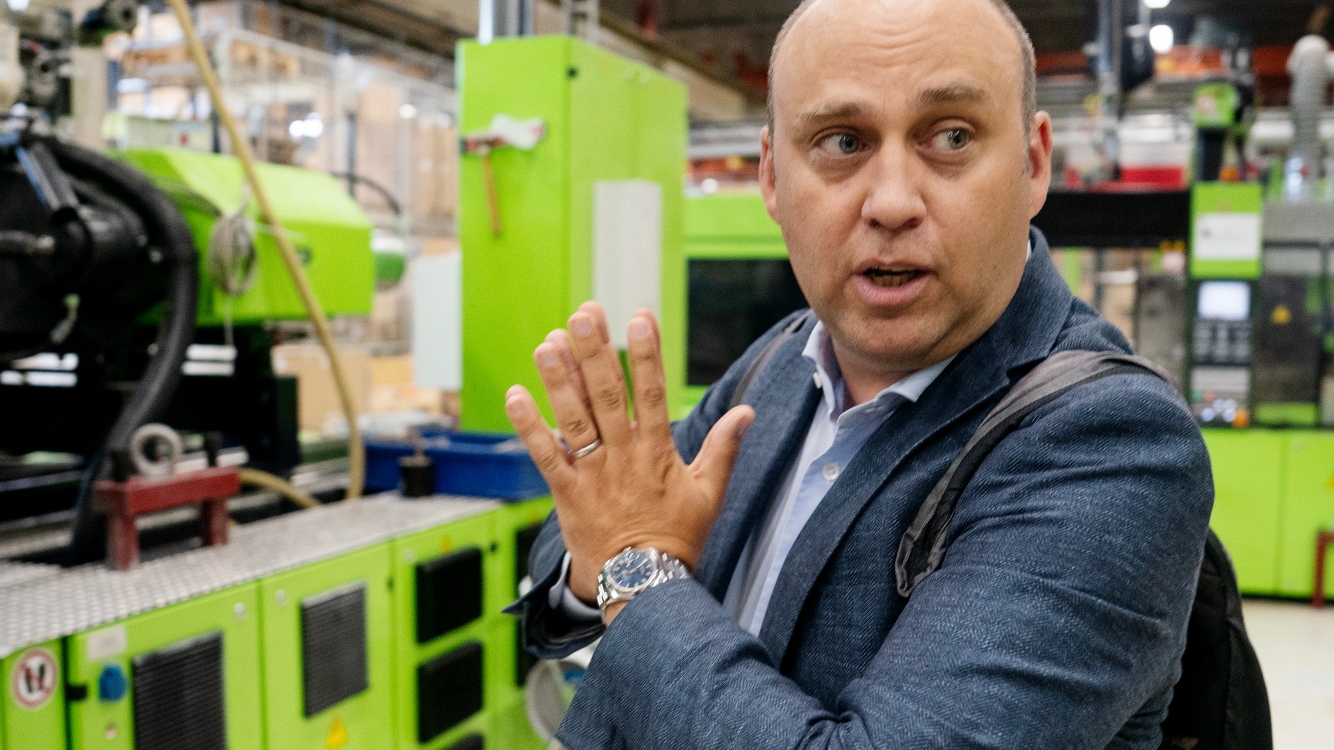 En man i mörkblå jacka gestikulerar och talar i en industriell miljö omgiven av grön maskinutrustning.
