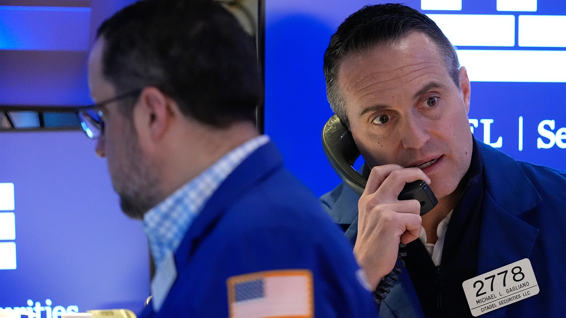 Två män på börsgolv med blå bakgrund, en av dem håller i en telefonheadsetBörsmäklare på Wall Street.