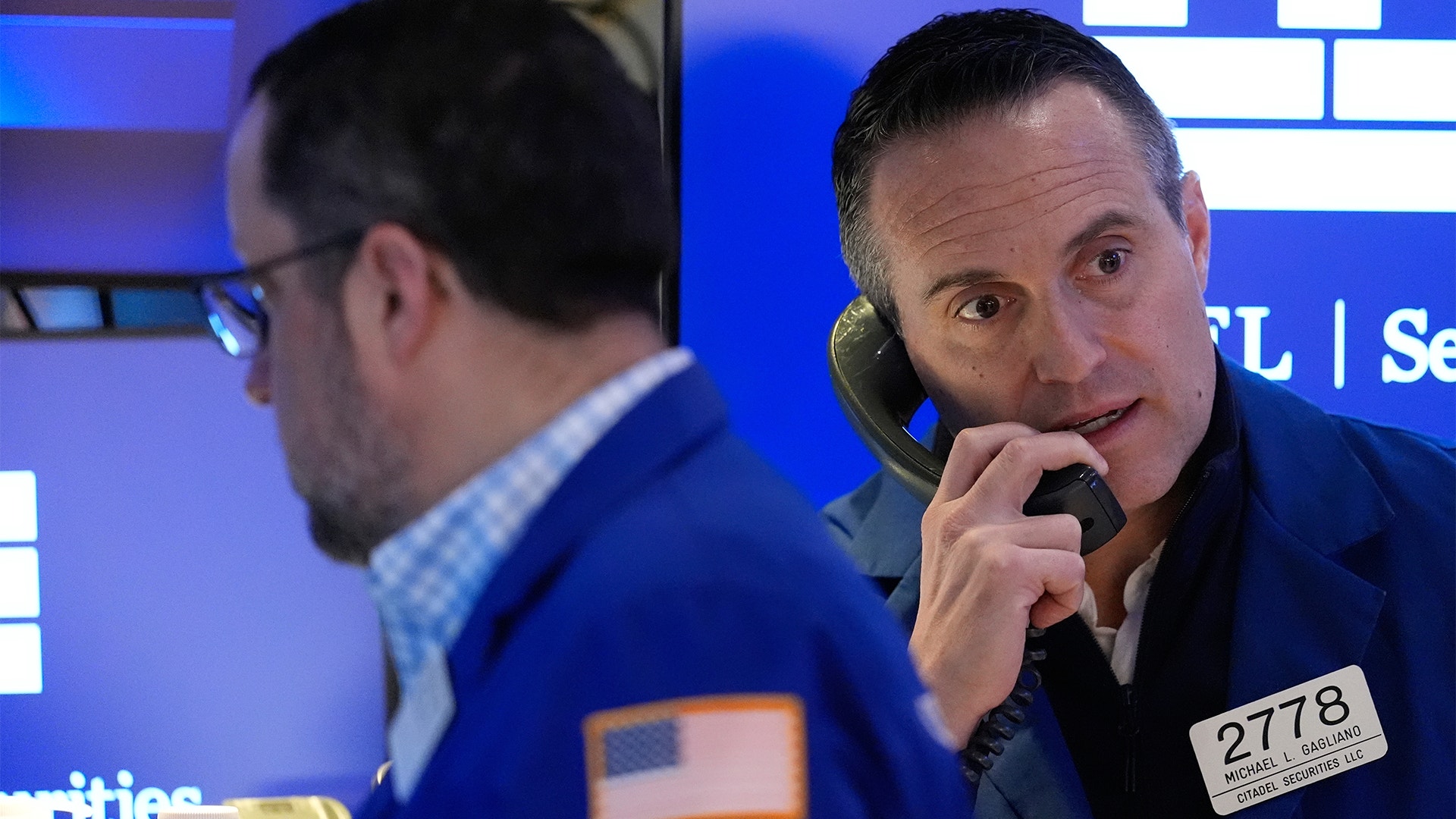 Två män på börsgolv med blå bakgrund, en av dem håller i en telefonheadsetBörsmäklare på Wall Street.
