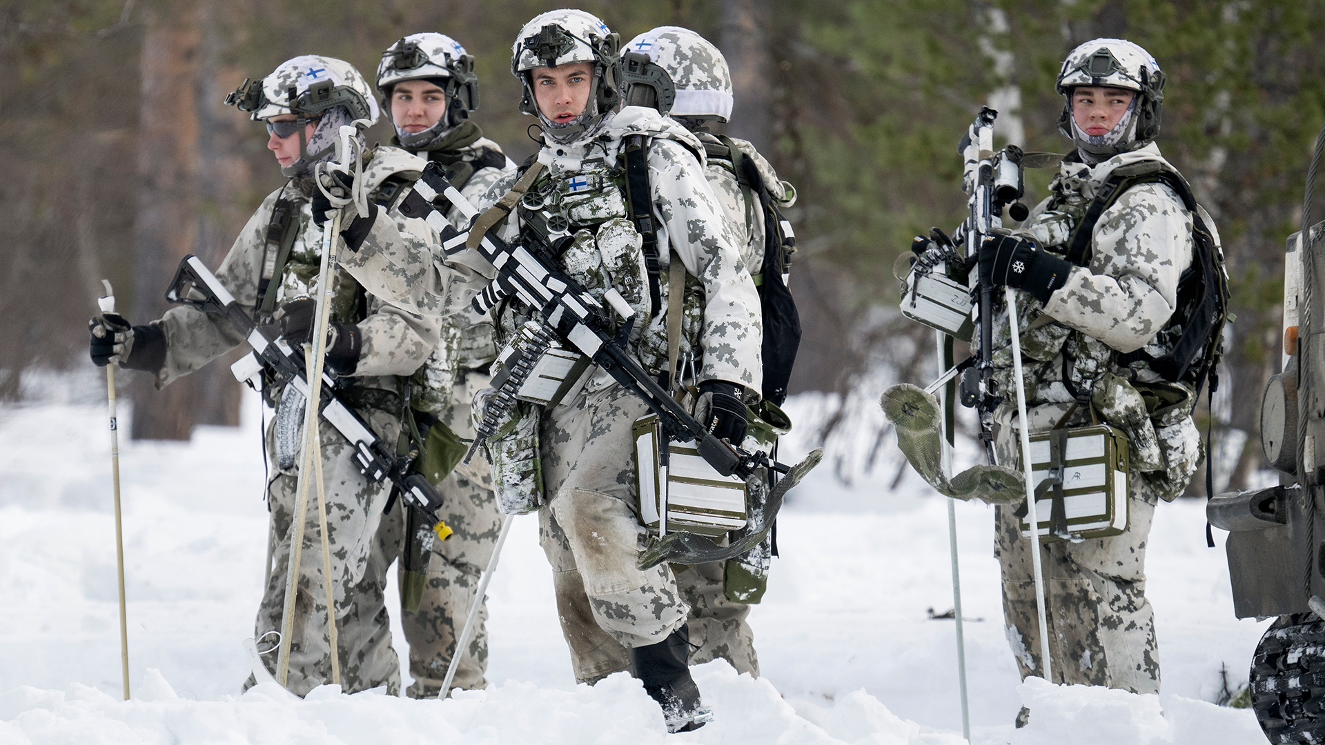 En grupp med militärer från det svenska försvaret står i skogen. De bär vapen och har uniformer på sig. Det är vinter och snö på marken.