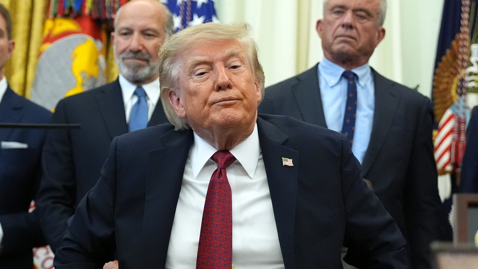 Donald Trump i mörk kostym med röd slips, omgiven av två män i blå kostymer i en officiell miljö med flaggor i bakgrunden.