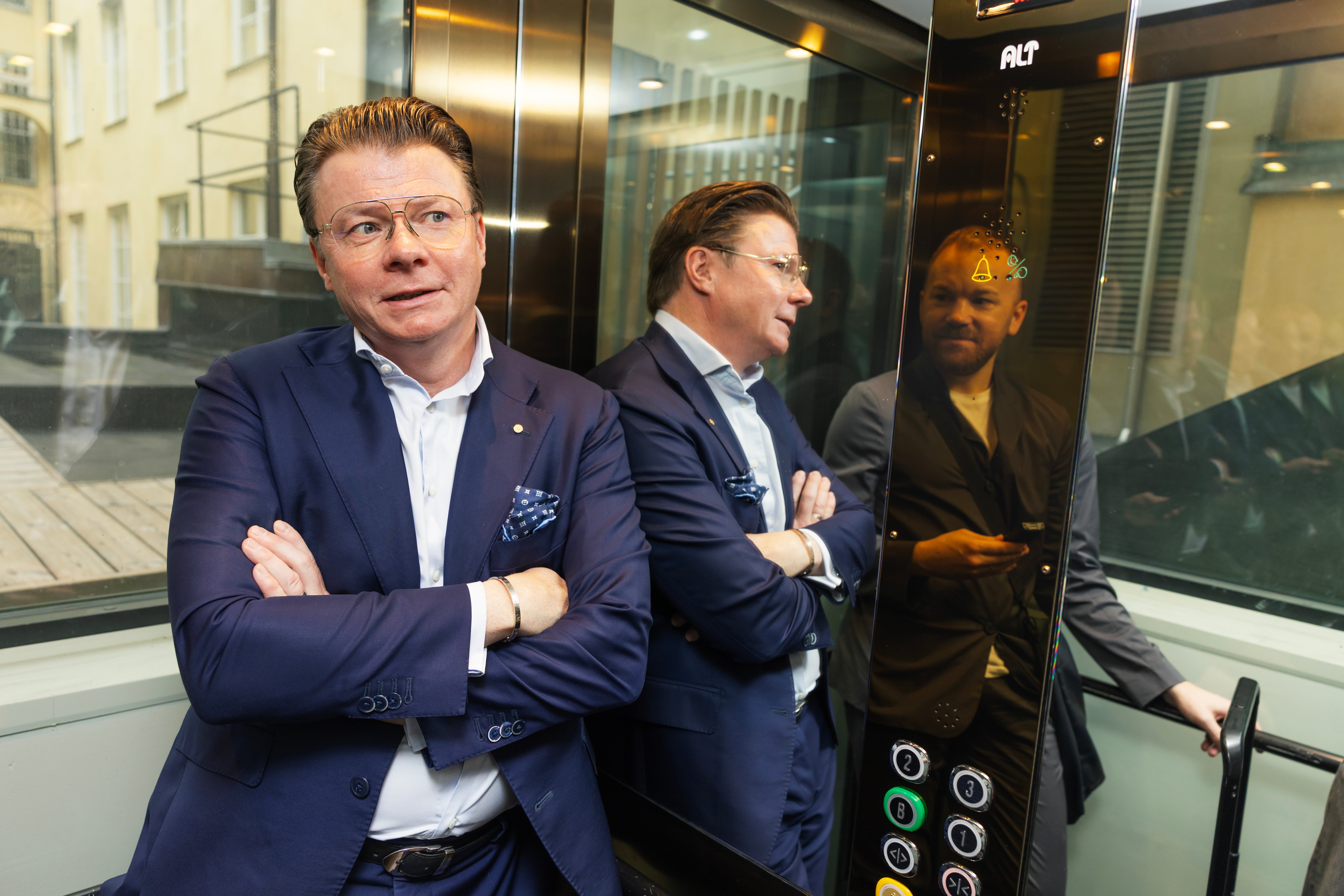 En man i kostym står med armarna i kors i en glashiss, med en annan person synlig i bakgrunden.