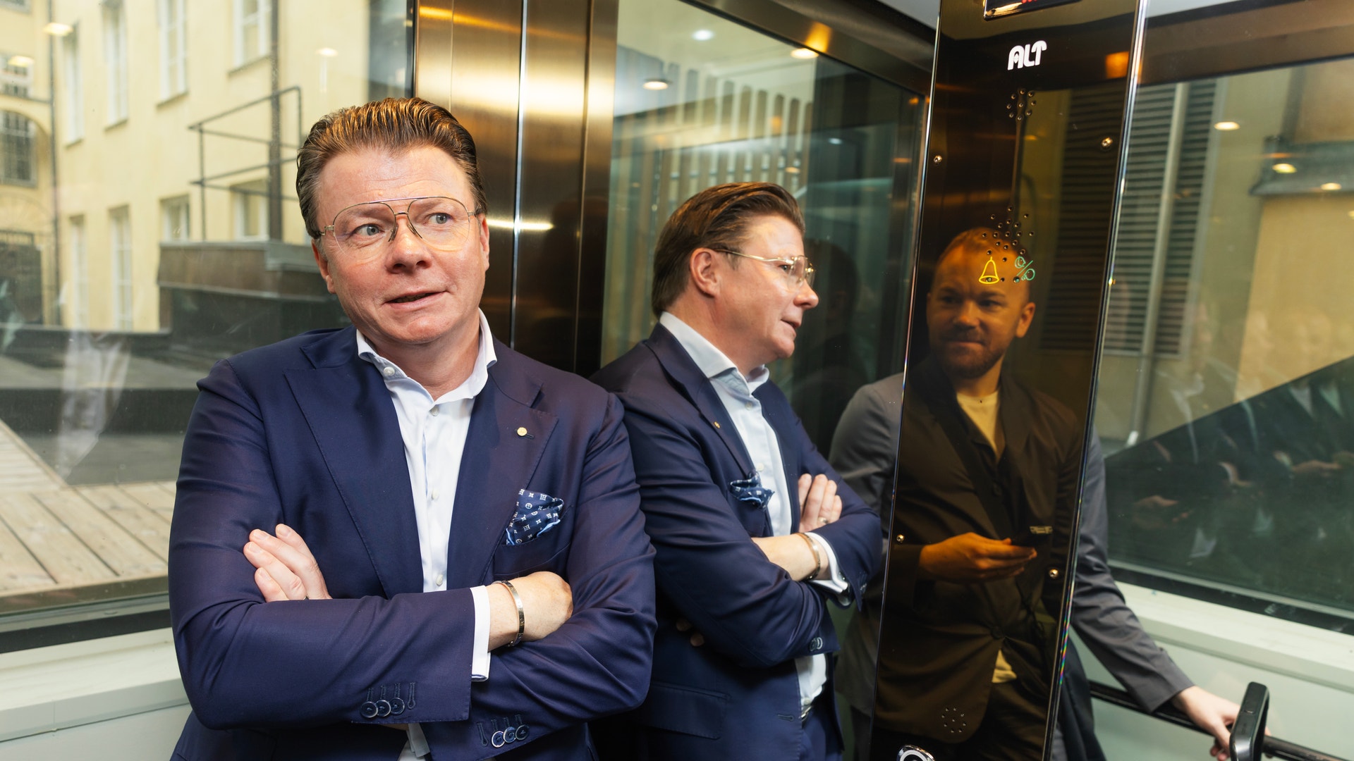 En man i kostym står med armarna i kors i en glashiss, med en annan person synlig i bakgrunden.