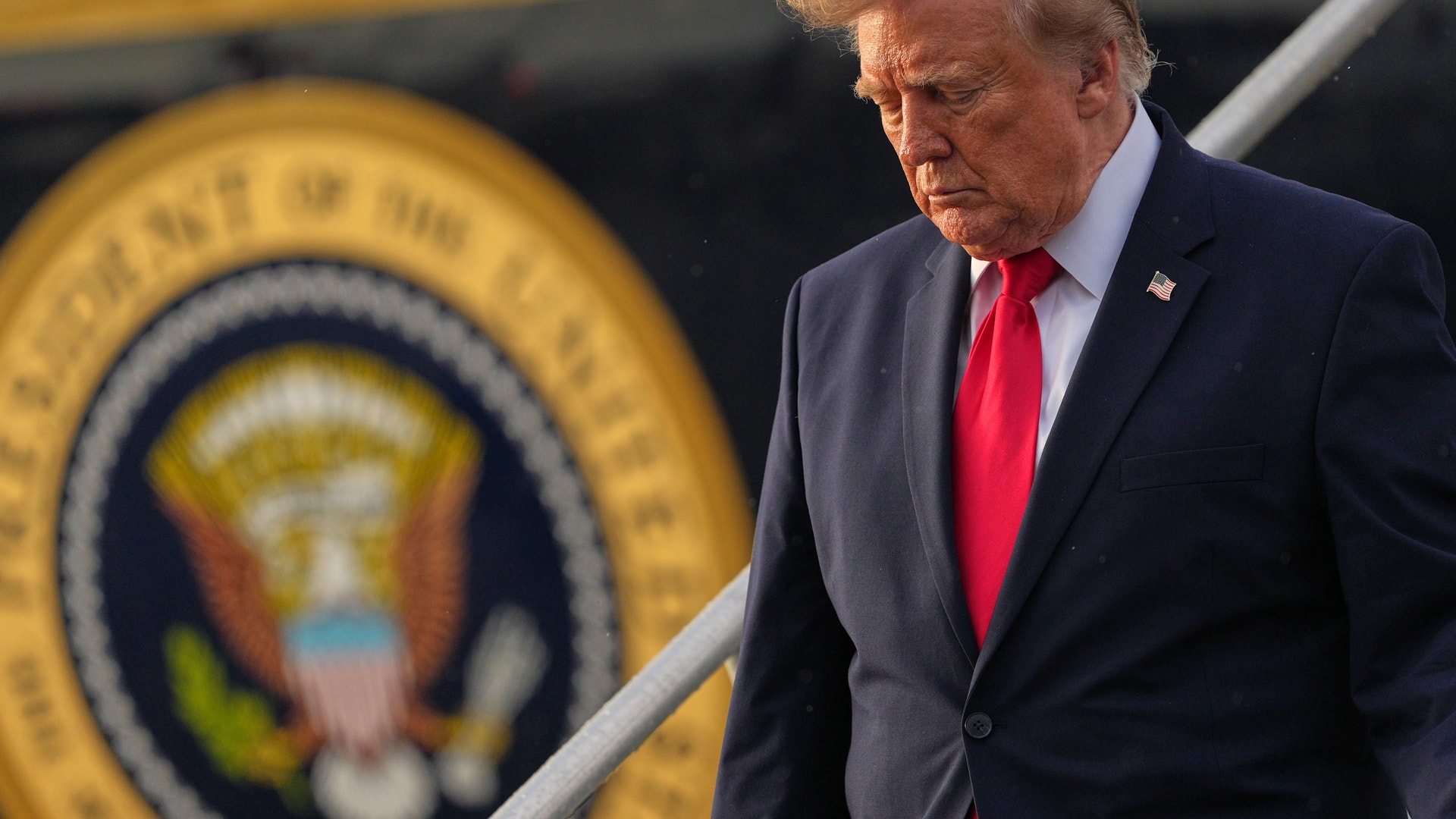 En äldre man i mörk kostym och röd slips går nedför en trappa med blicken riktad nedåt. Bakom honom syns USA:s presidentsigill.