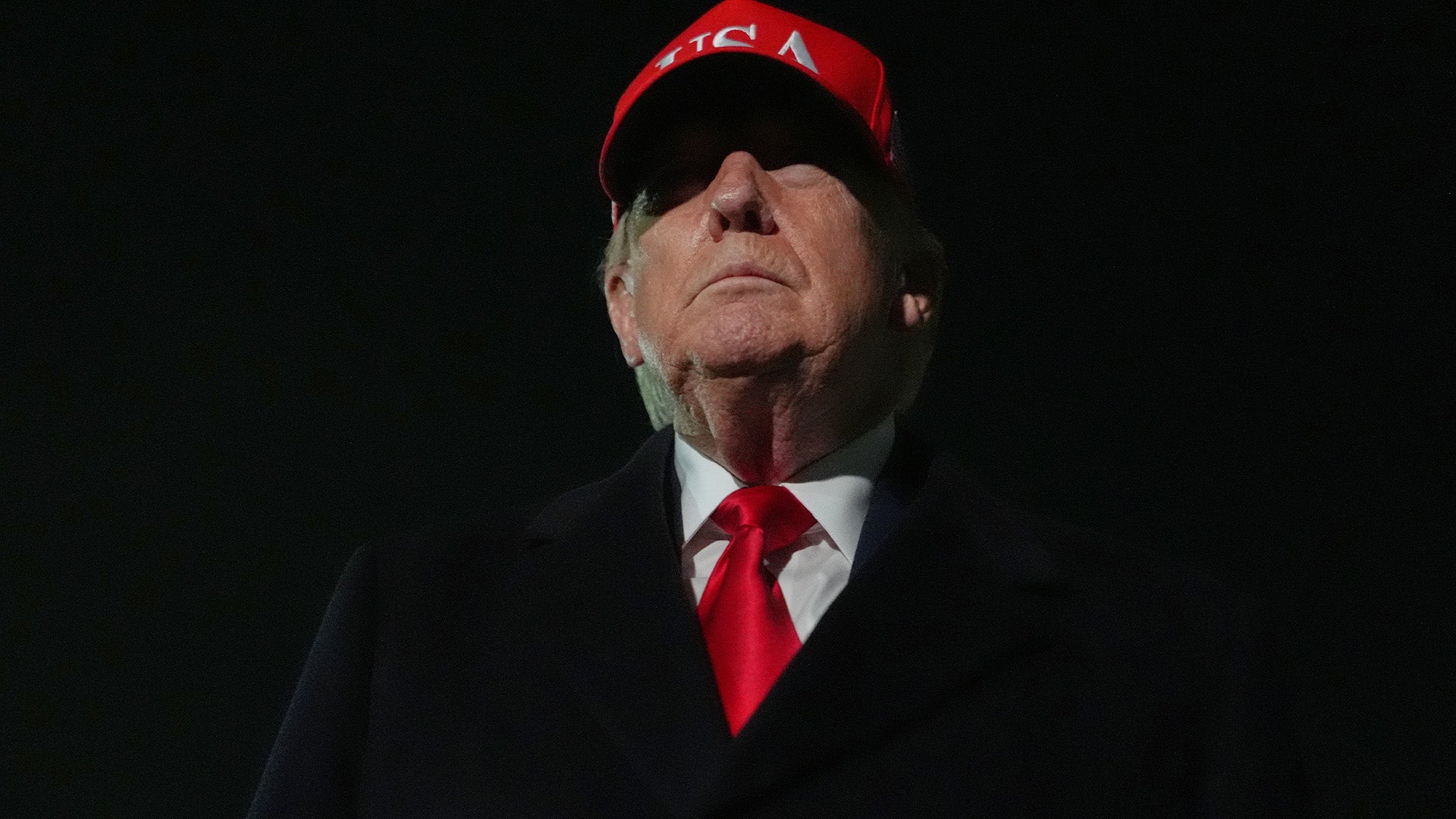 Porträtt av en man i röd keps och formell kostym mot mörk bakgrund.