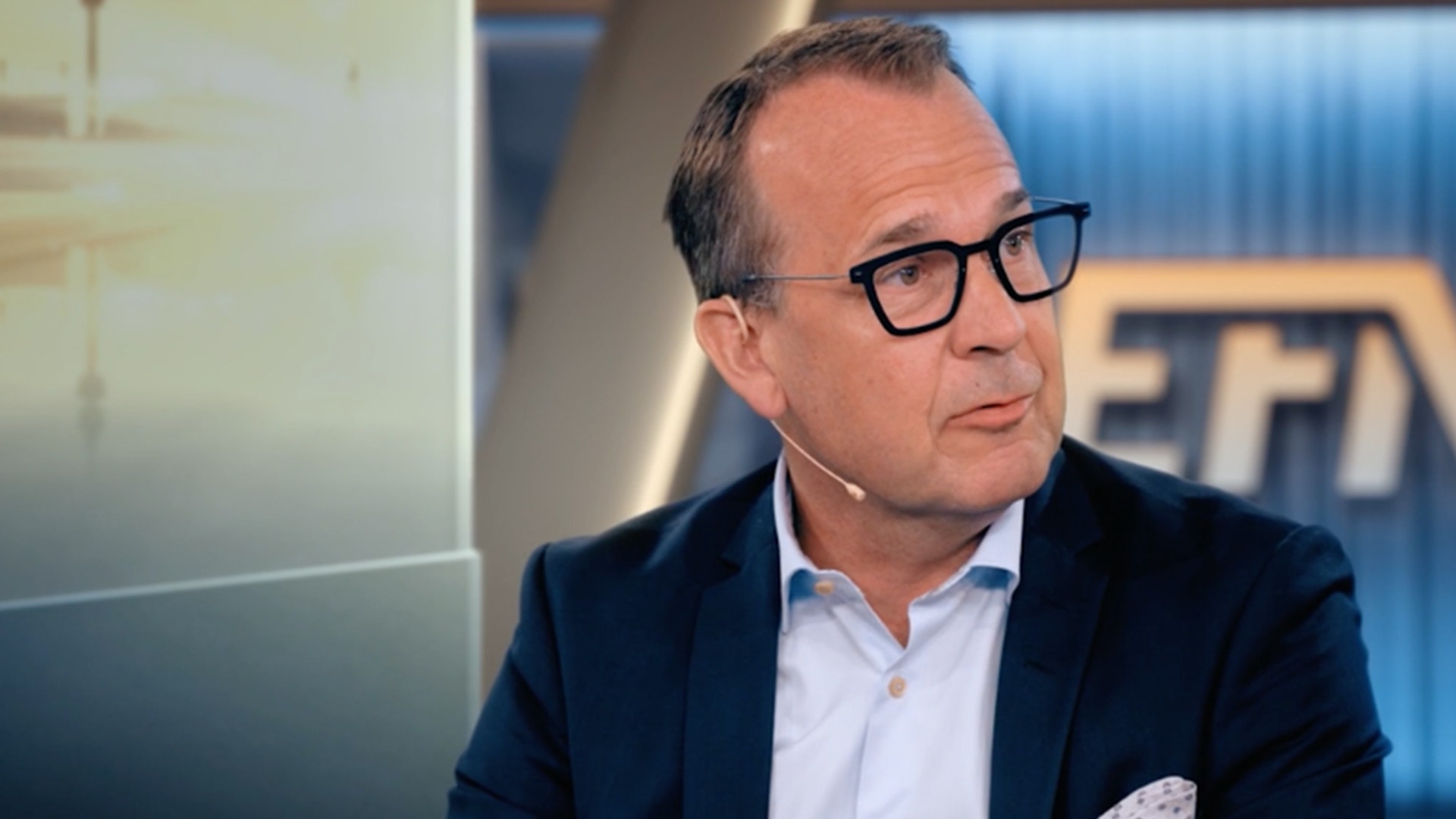 Porträttfoto av en man med glasögon och mörk blazer i en professionell studiomiljö.