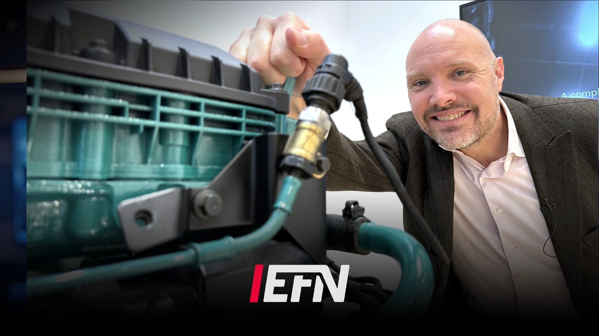 En man i kostym presenterar en bilkomponent framför en bilmotor. EFN-logga visas i bilden.